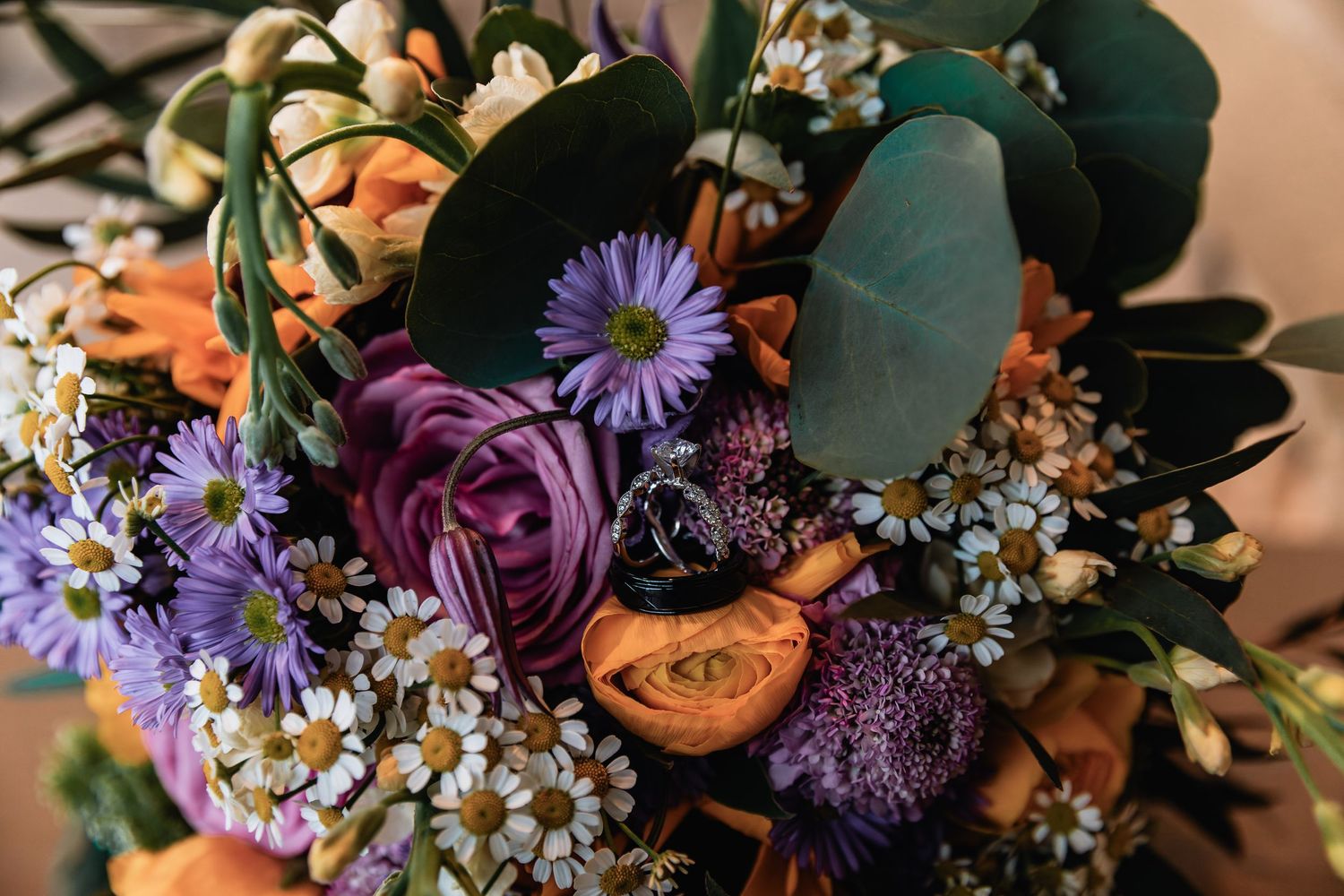 Close-up floral arrangement with purple roses, orange tulips, blue asters and eucalyptus leaves in vibrant colors.