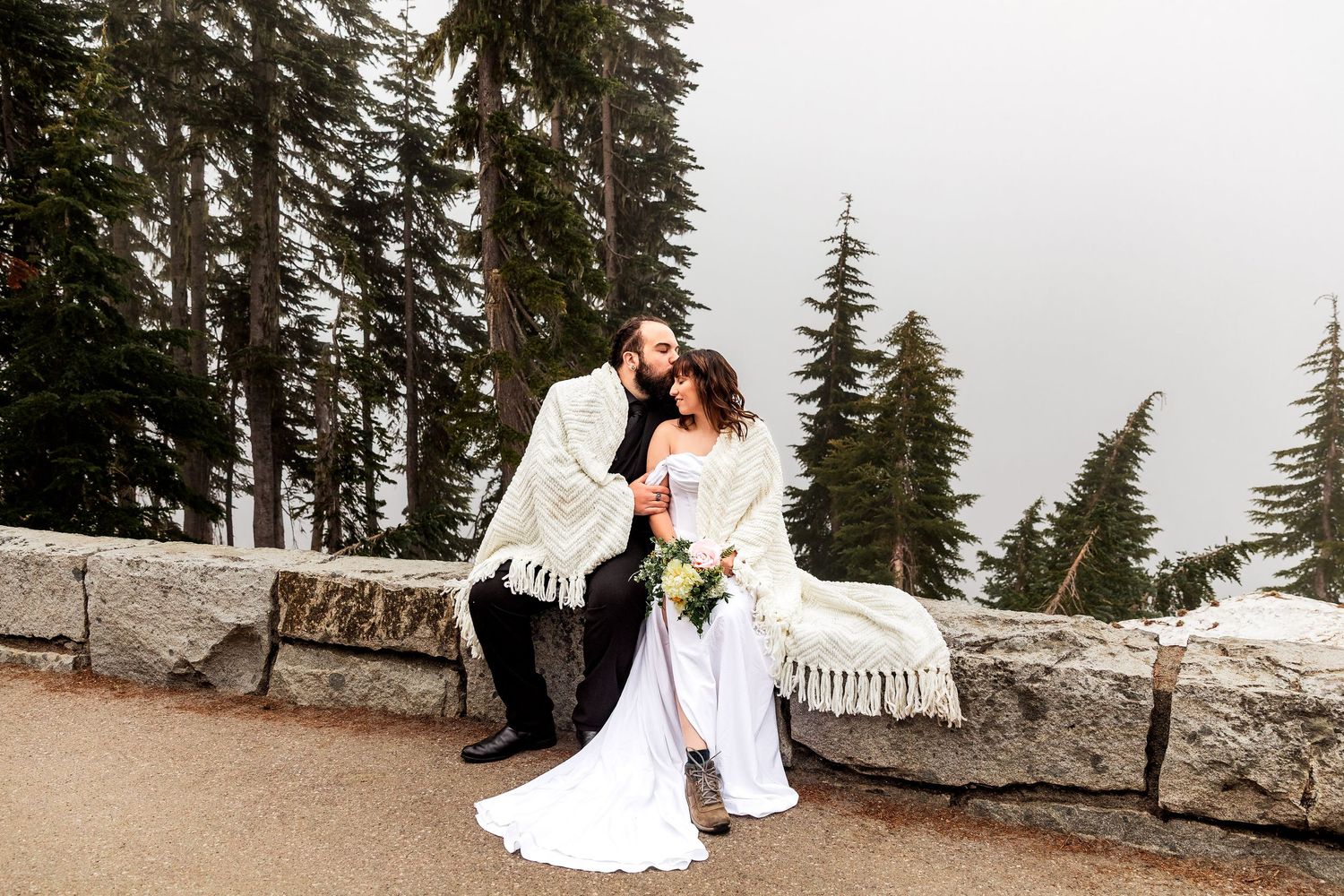 A couple in wedding attire sits together on a stone bench surrounded by evergreen trees and patches of snow.