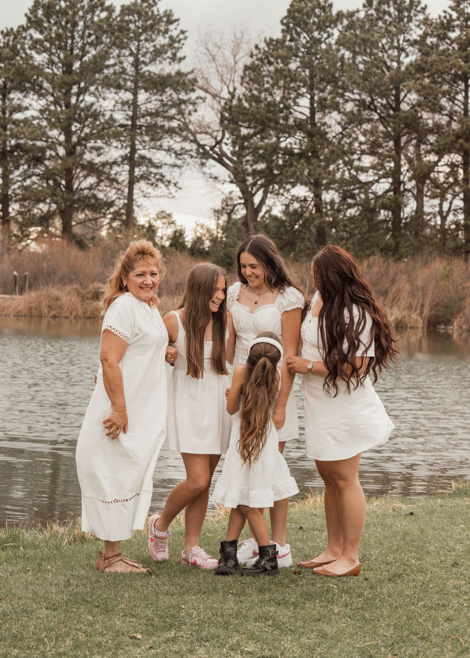Women in white dresses laugh together on grass near a lake during a photo session.