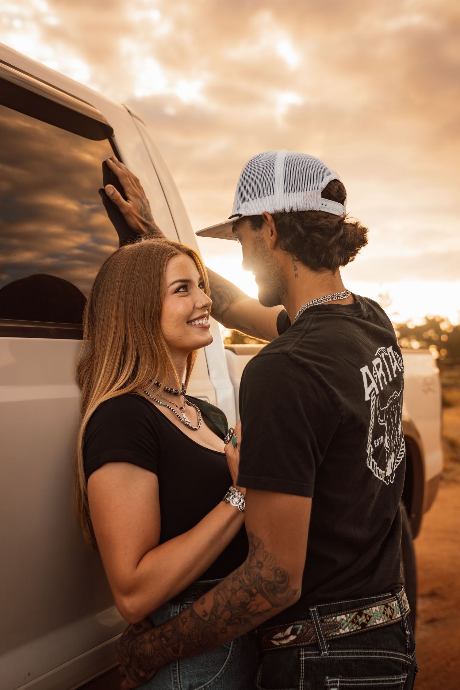 A smiling couple leans against a van at sunset, the man in a snapback cap and the woman in a black top with tattoos.