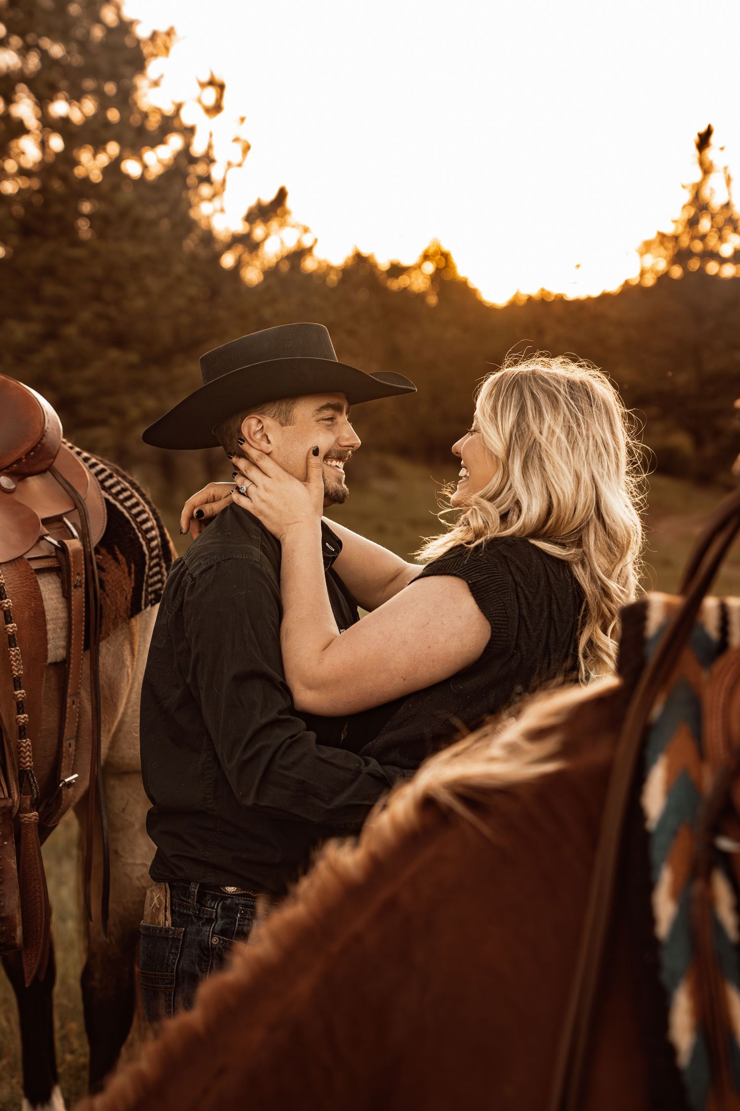 A couple in western attire shares a romantic moment at golden hour, with horses nearby and trees in the background.