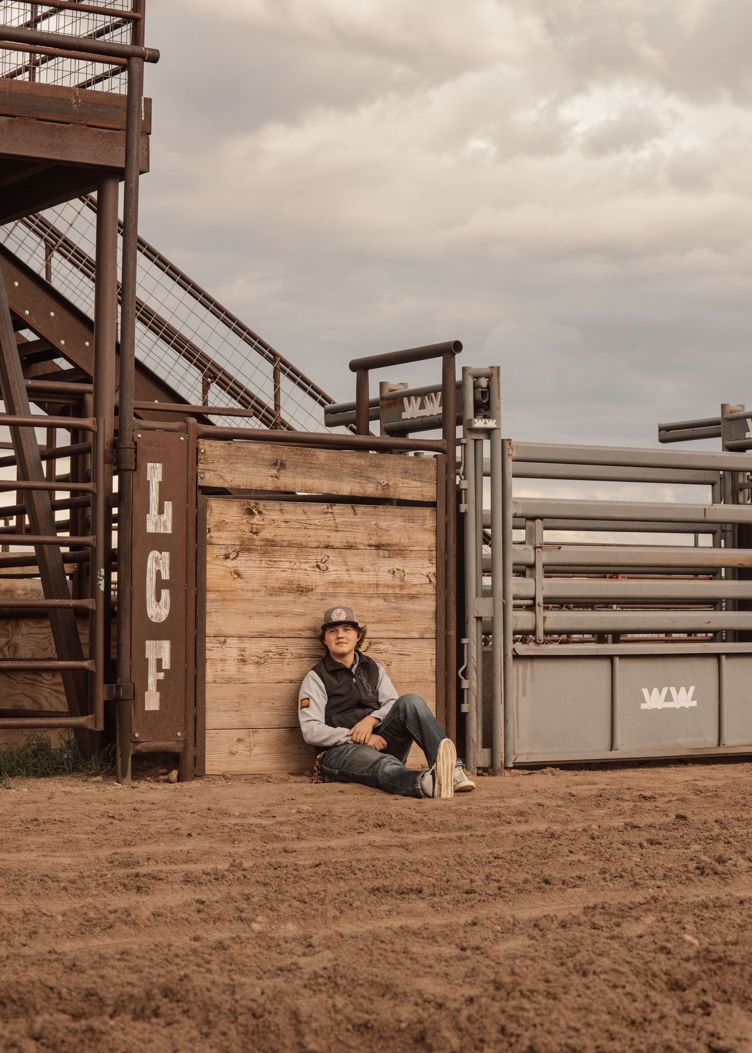Person in western clothing sits near wooden rodeo gates in a dirt arena.