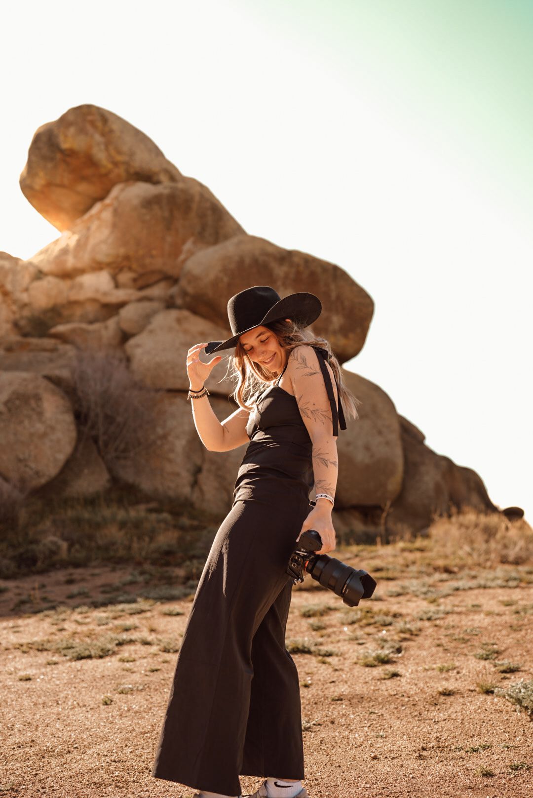 Woman in black wide-leg jumpsuit and hat holds camera in desert landscape with large boulders at golden hour.