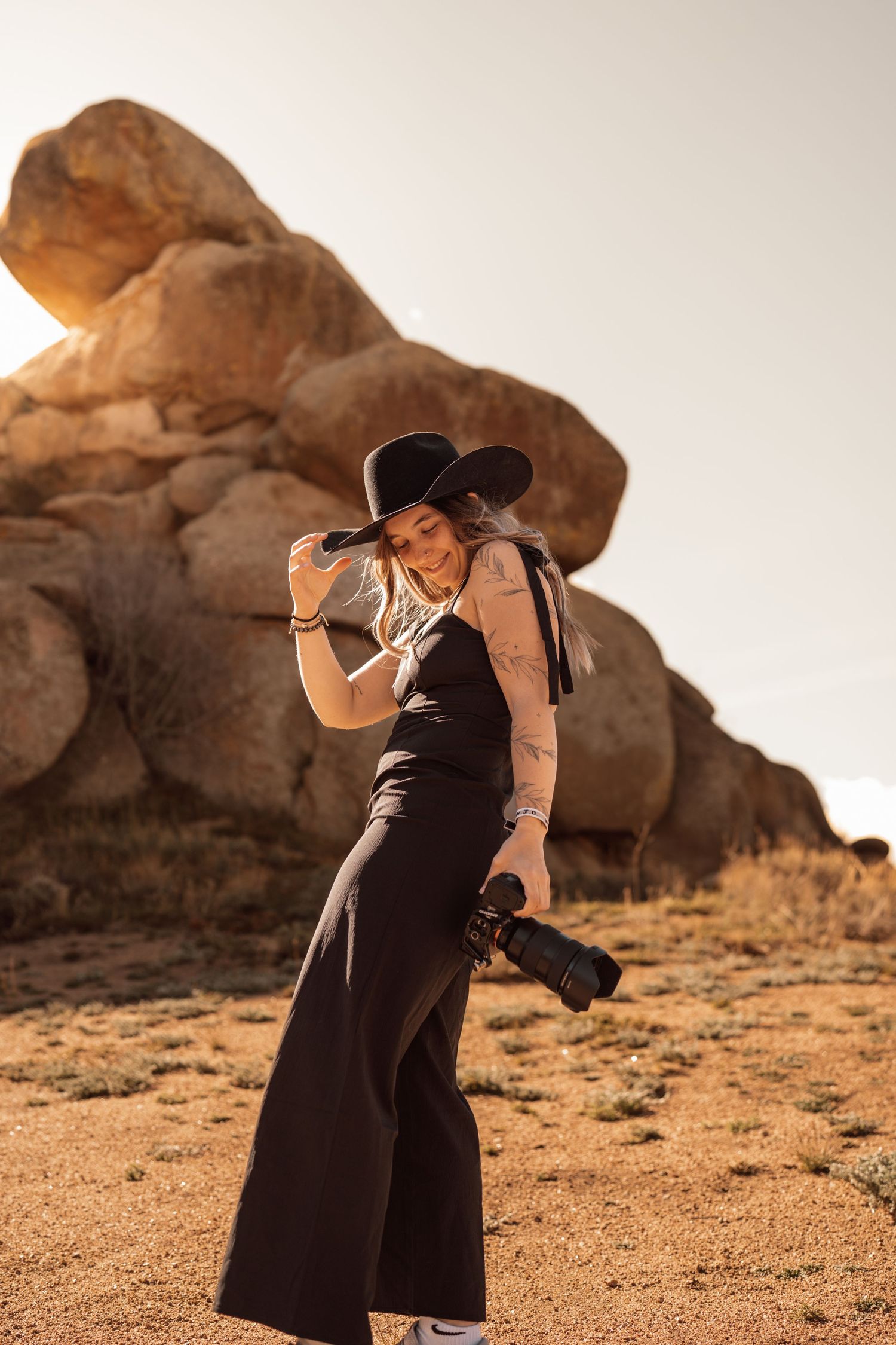 Woman in black wide-brim hat and jumpsuit holds camera, smiling in desert landscape with golden rock formations.