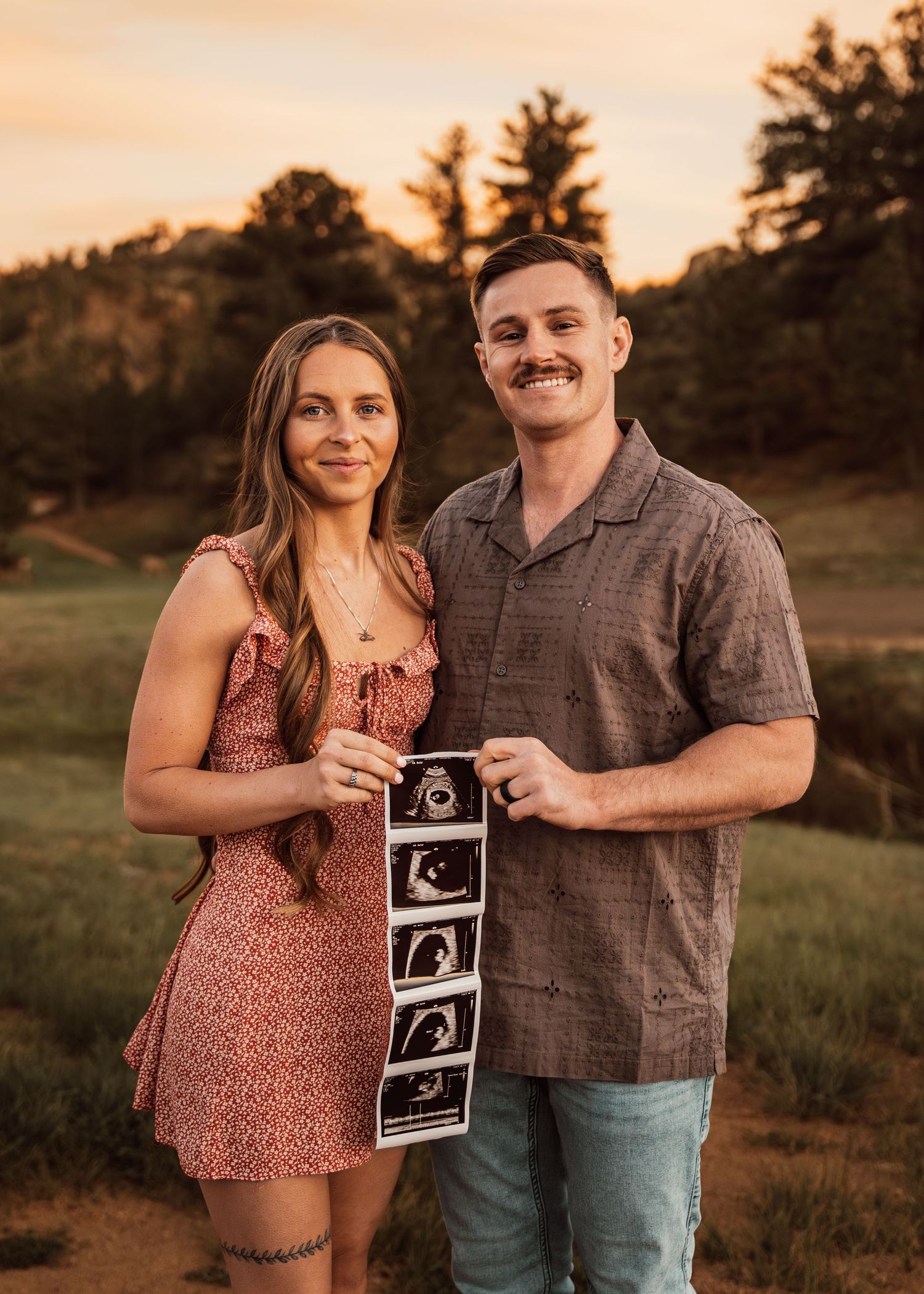 Smiling couple holds ultrasound photos outdoors at golden hour, announcing their pregnancy in a scenic field.