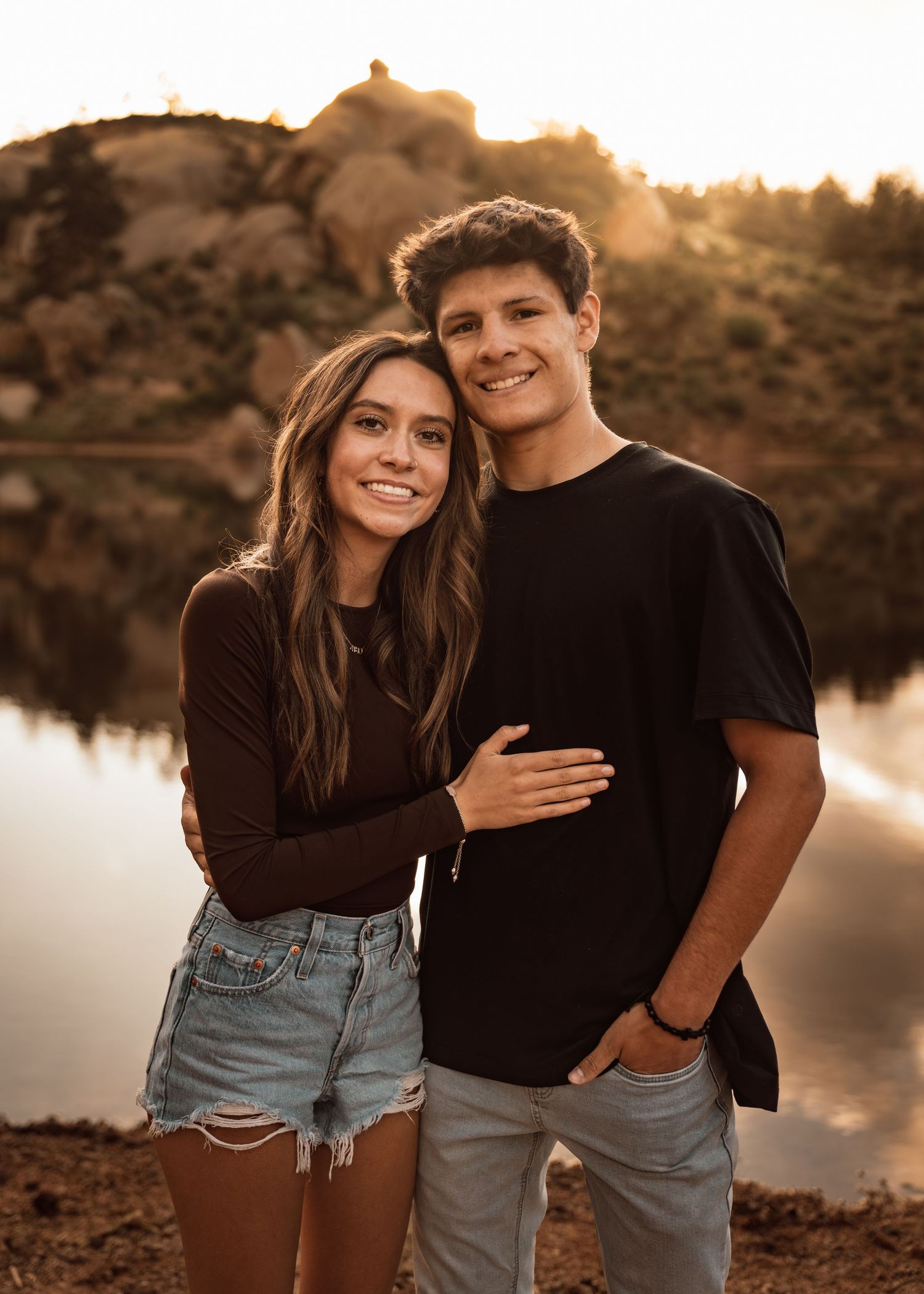 A young couple poses together near a lake at golden hour, smiling warmly in casual black tops and denim.