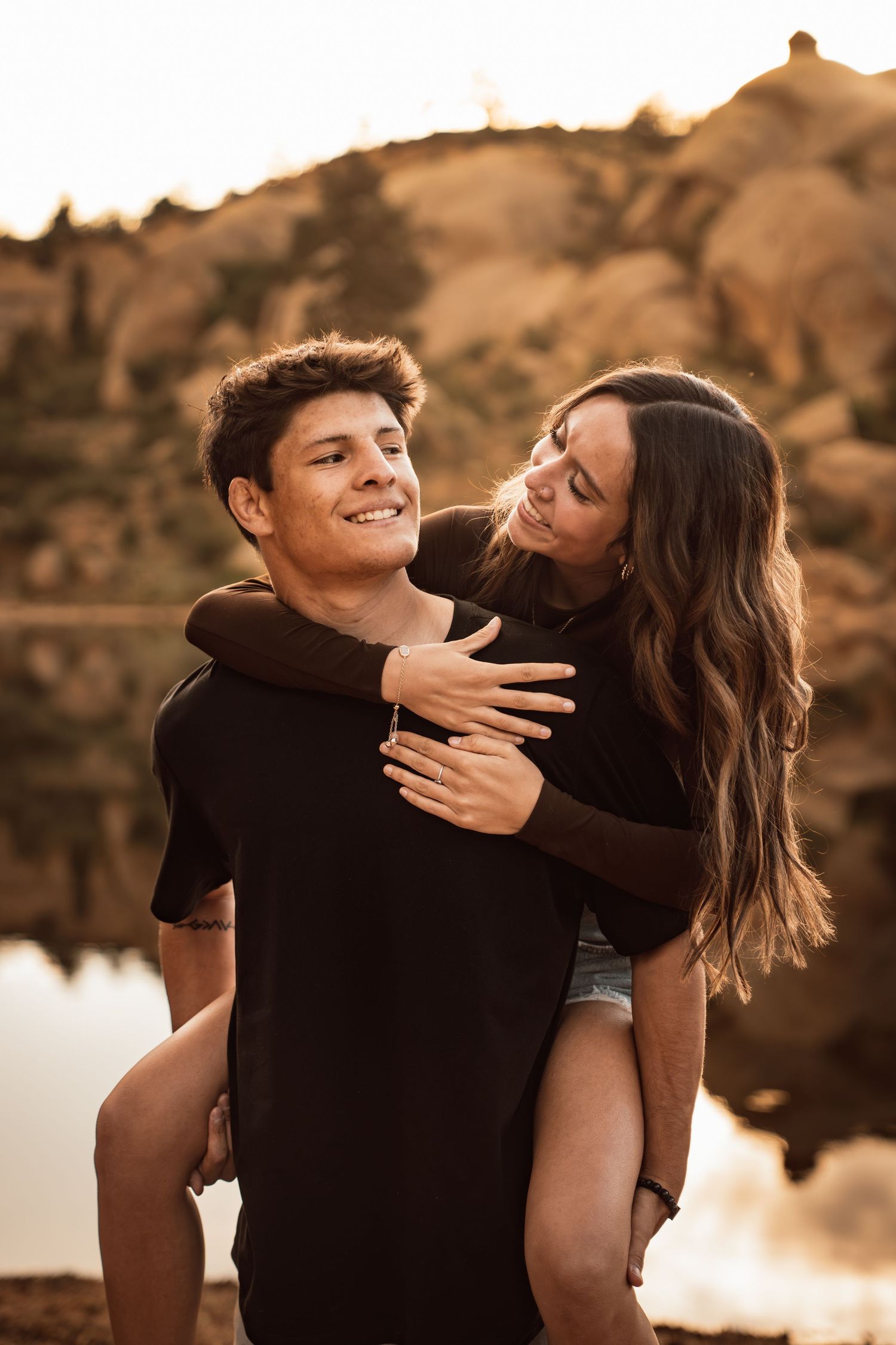 A young couple shares a joyful piggyback ride outdoors at golden hour near a scenic rocky lake.