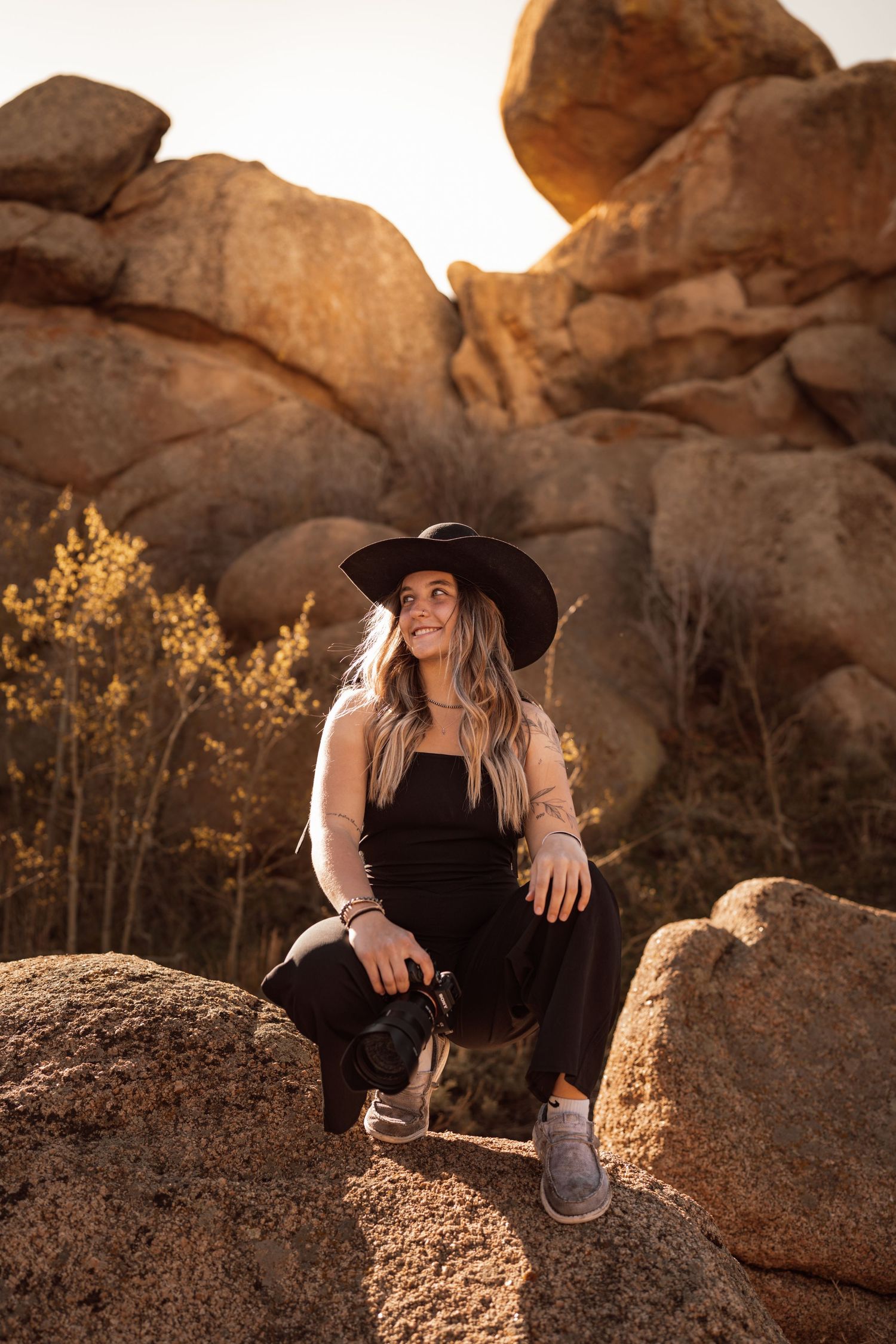 Woman in black outfit and wide-brim hat sits on desert rocks holding camera, golden hour light on boulders behind her.