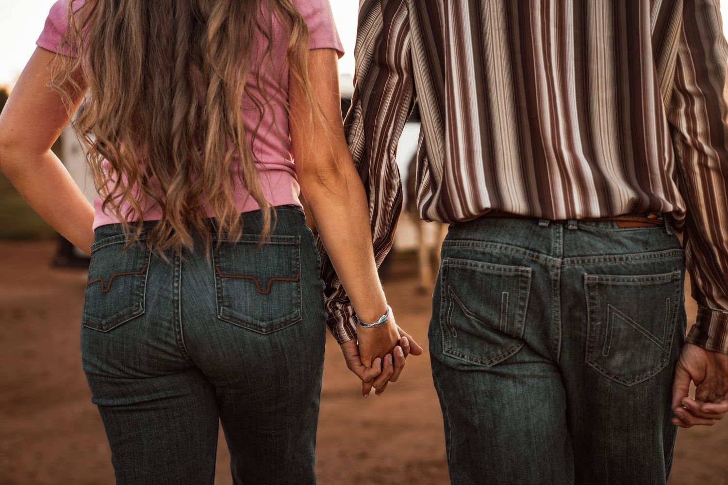 Two people holding hands from behind, wearing jeans and casual tops, standing outdoors in warm light.