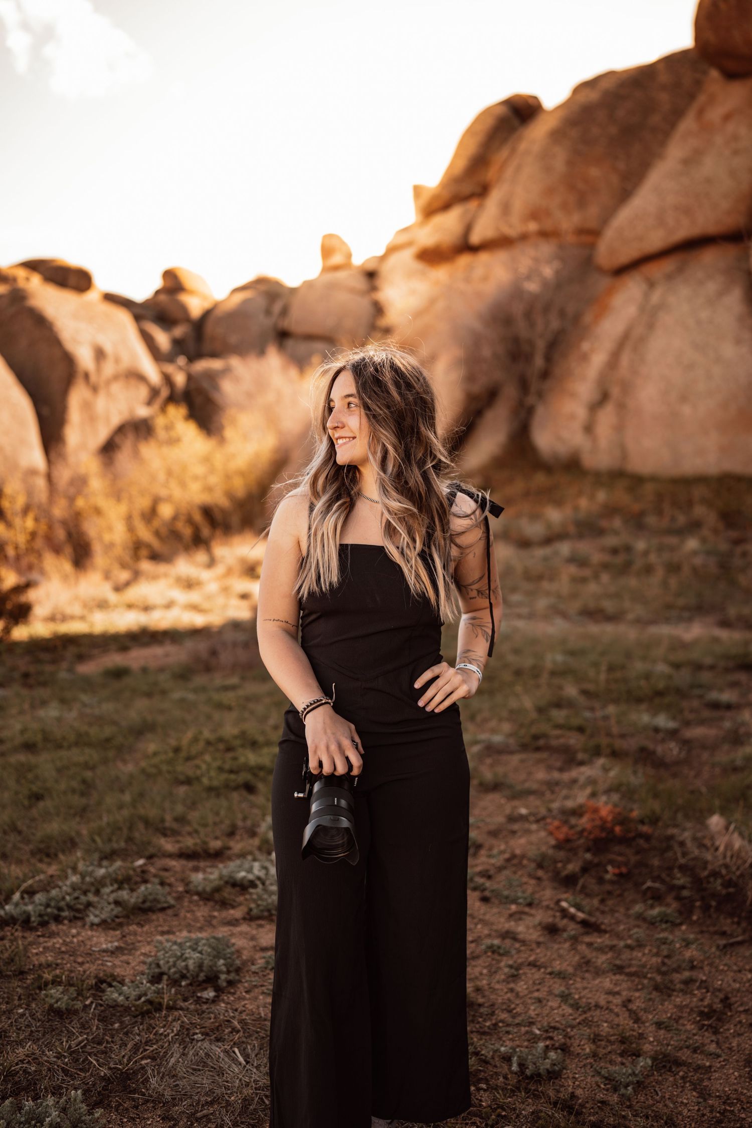 Woman in black jumpsuit holding camera smiling outdoors near large boulders at golden hour.