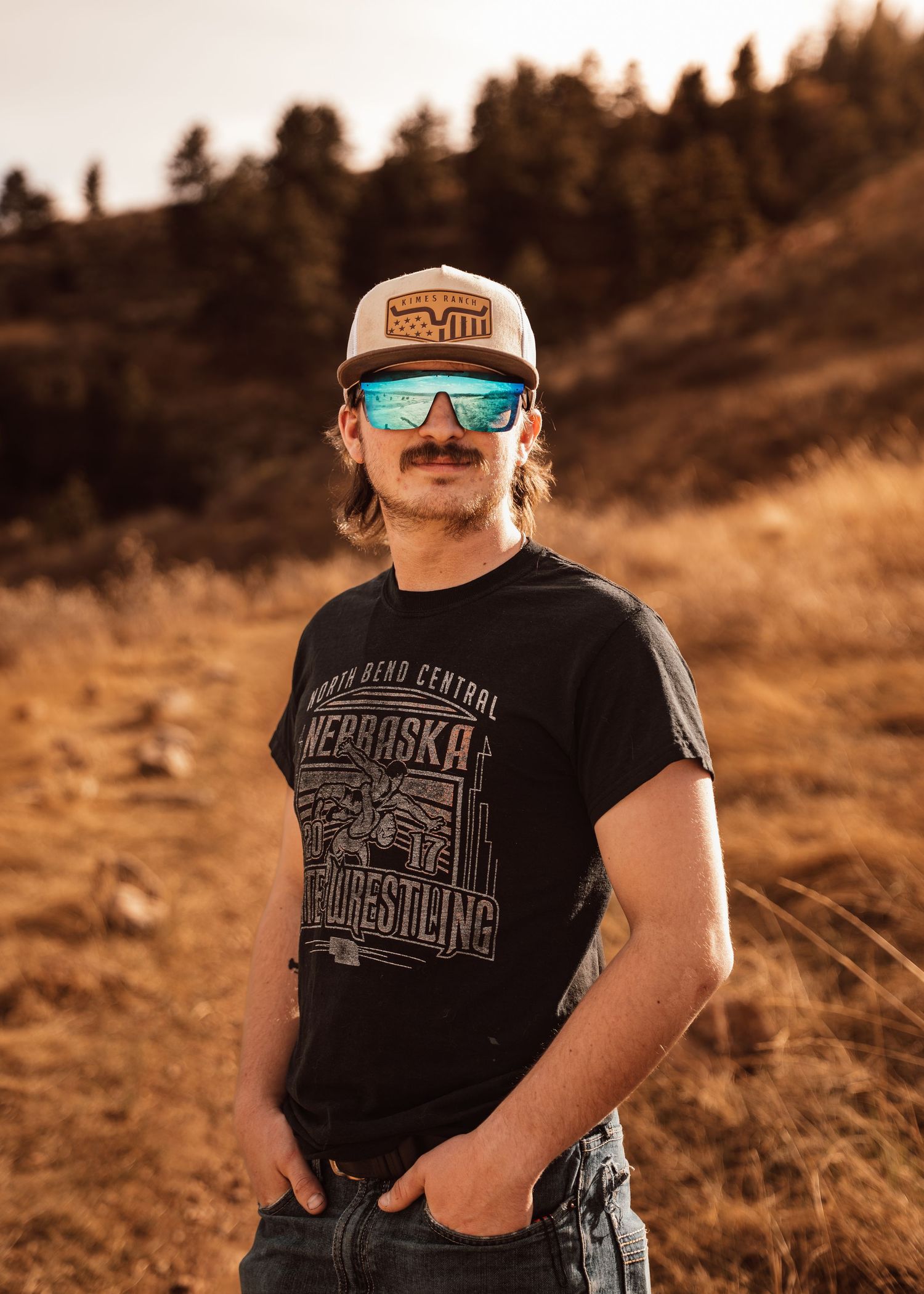 Young man with mustache wearing trucker hat, blue mirrored sunglasses, and Nebraska Wrestling t-shirt outdoors.