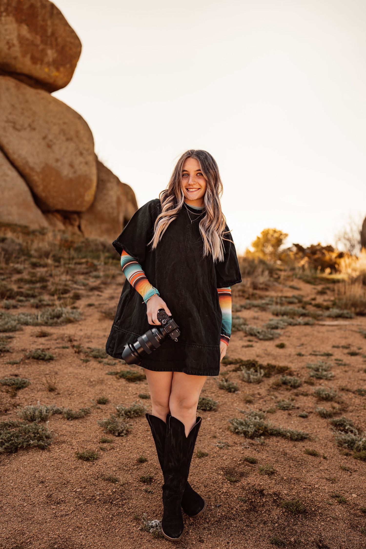 Woman in black dress and cowboy boots holds camera in desert landscape with large rocks at golden hour.
