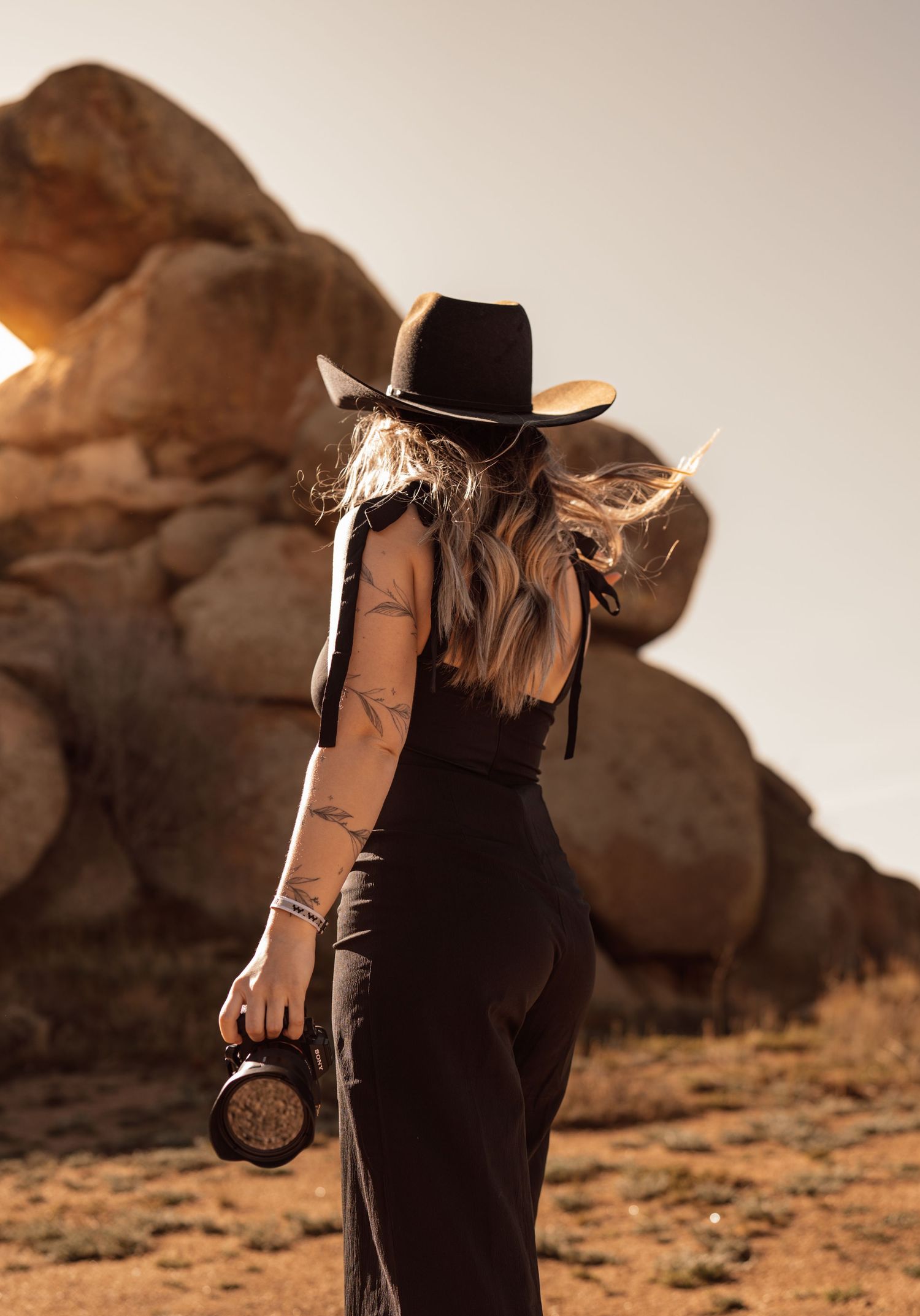 Woman in black outfit and cowboy hat holding a lantern in a desert landscape with large boulders at sunset.