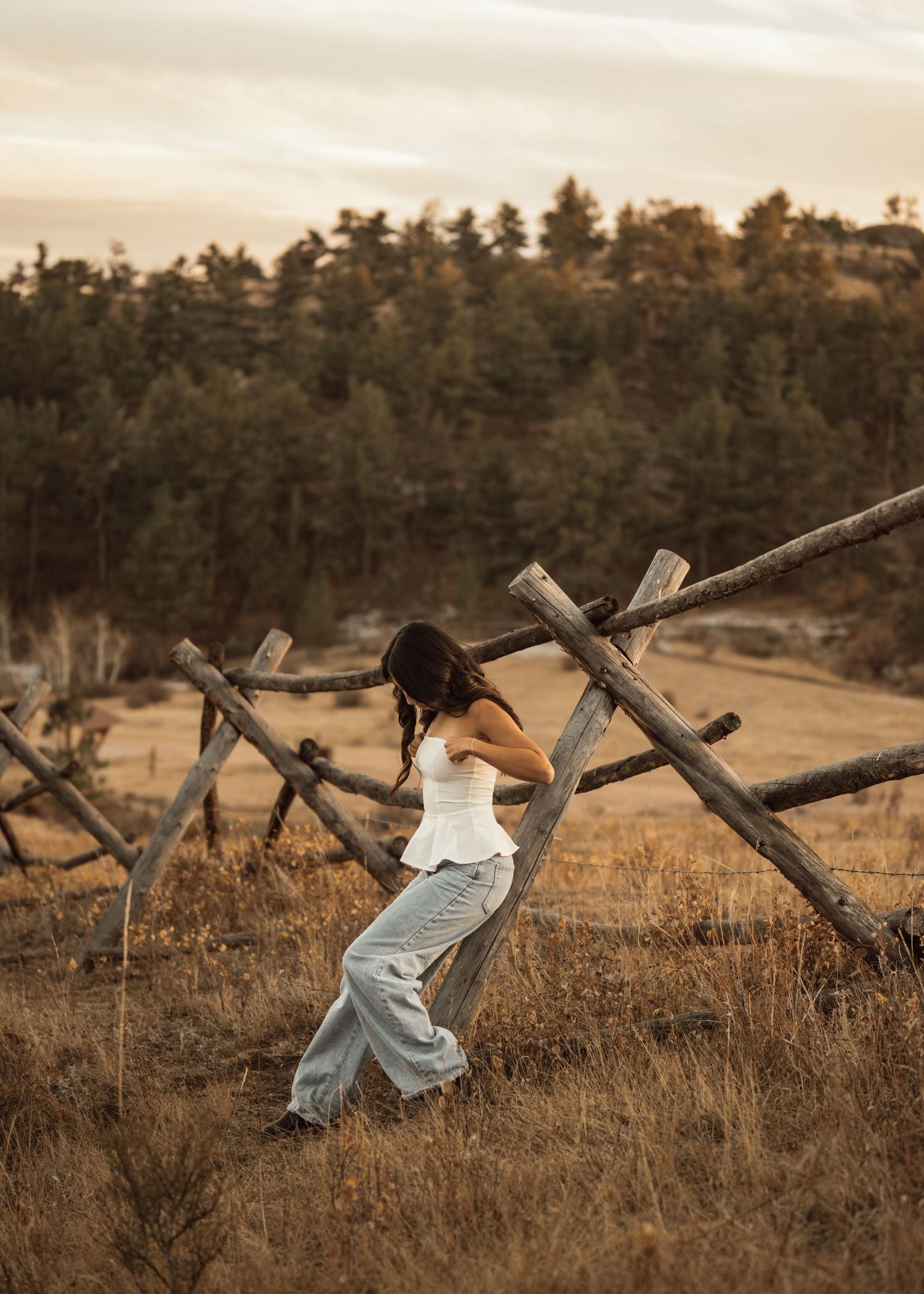 Woman in flowing dress poses dramatically near rustic wooden fence at sunset.