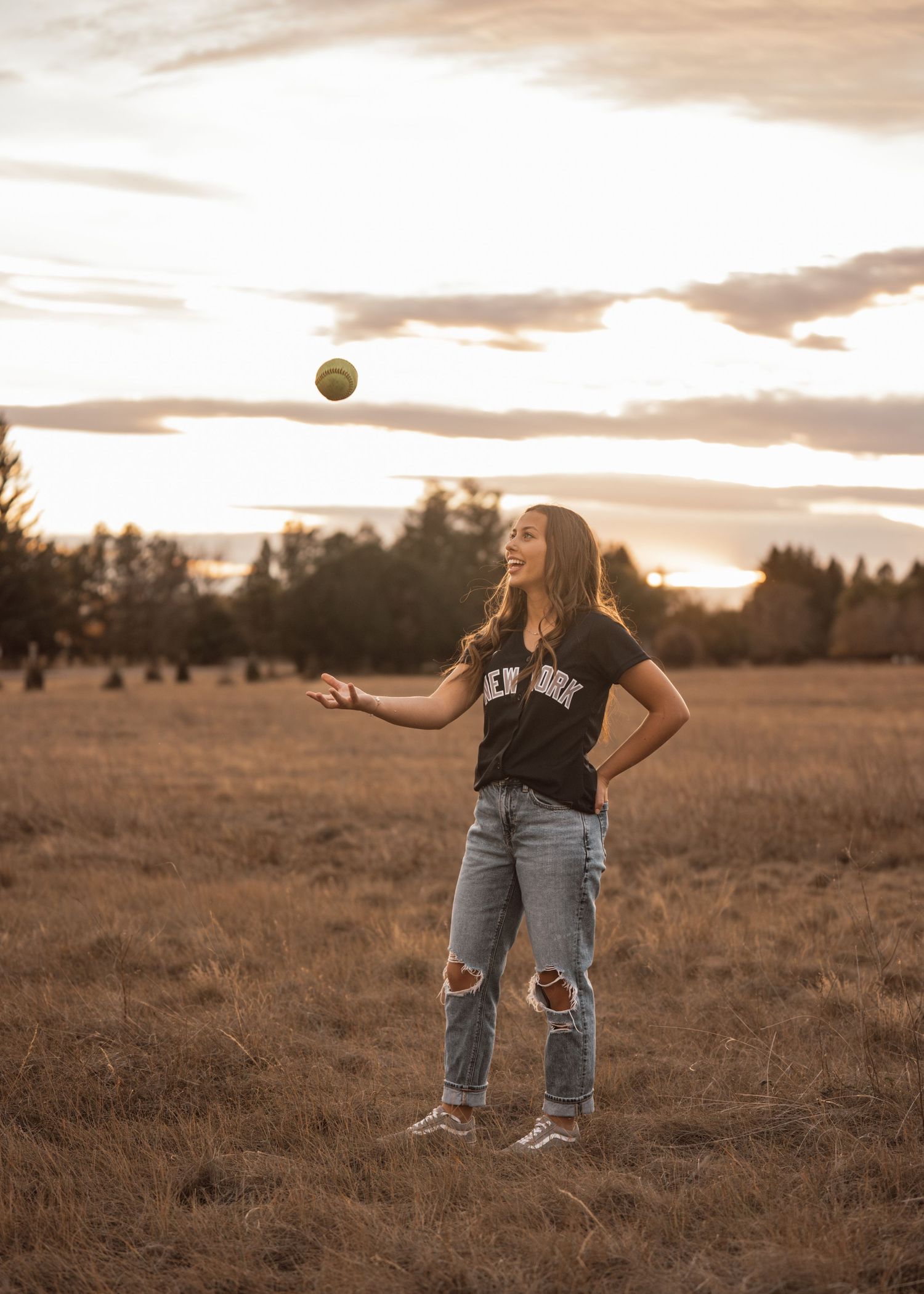 Person tosses softball in air while standing in golden field at sunset.