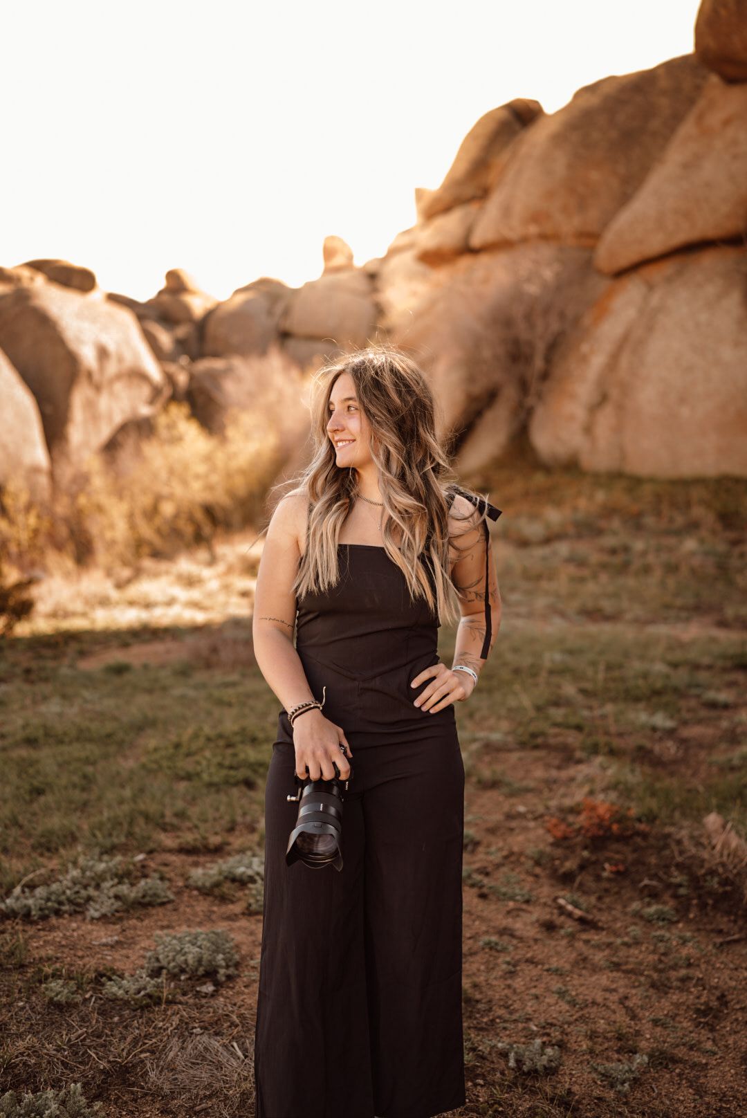 Woman in black jumpsuit holding camera smiling at golden hour near rocky landscape.