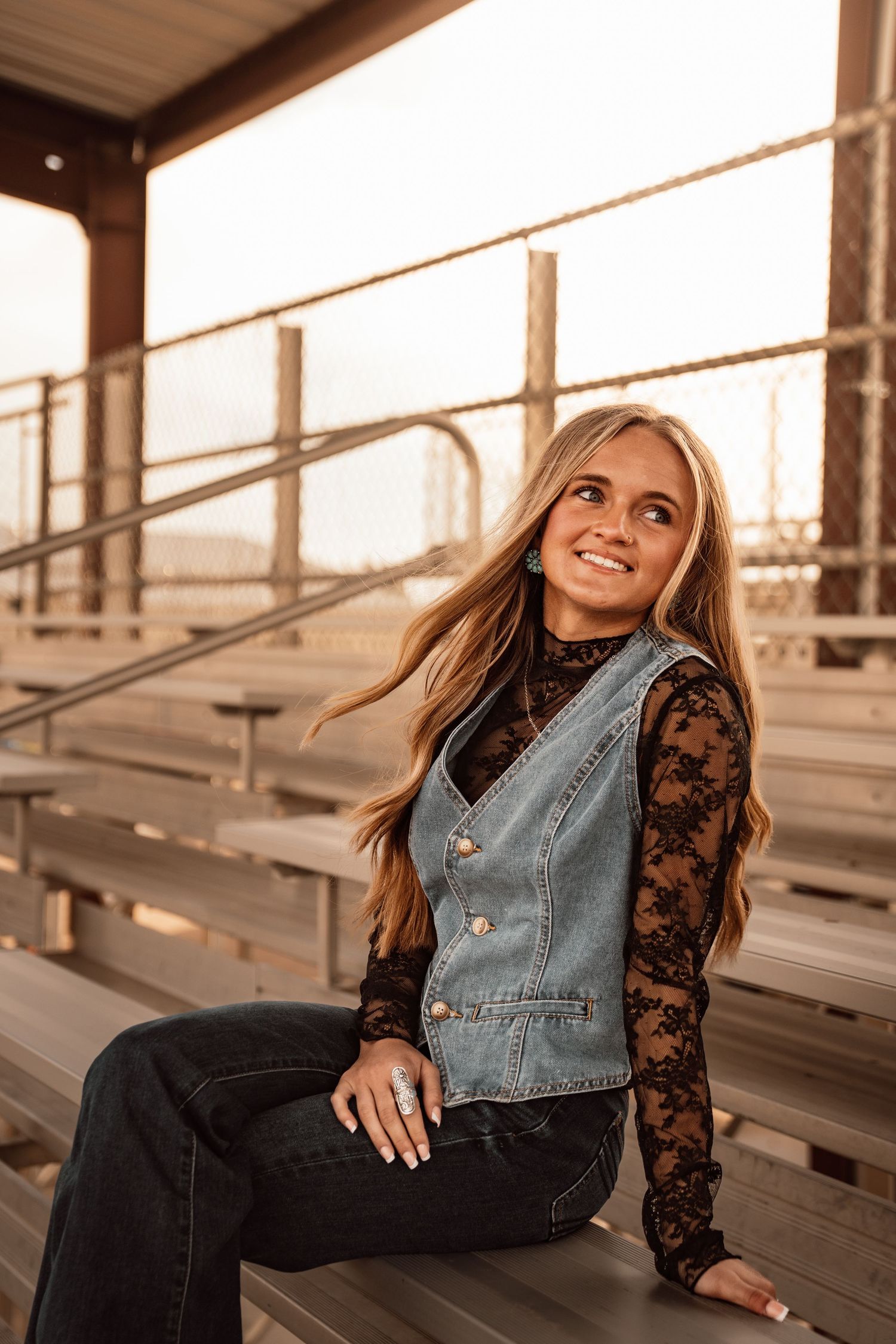 Young blonde woman smiling in denim vest and lace top, sitting on bleachers at golden hour.