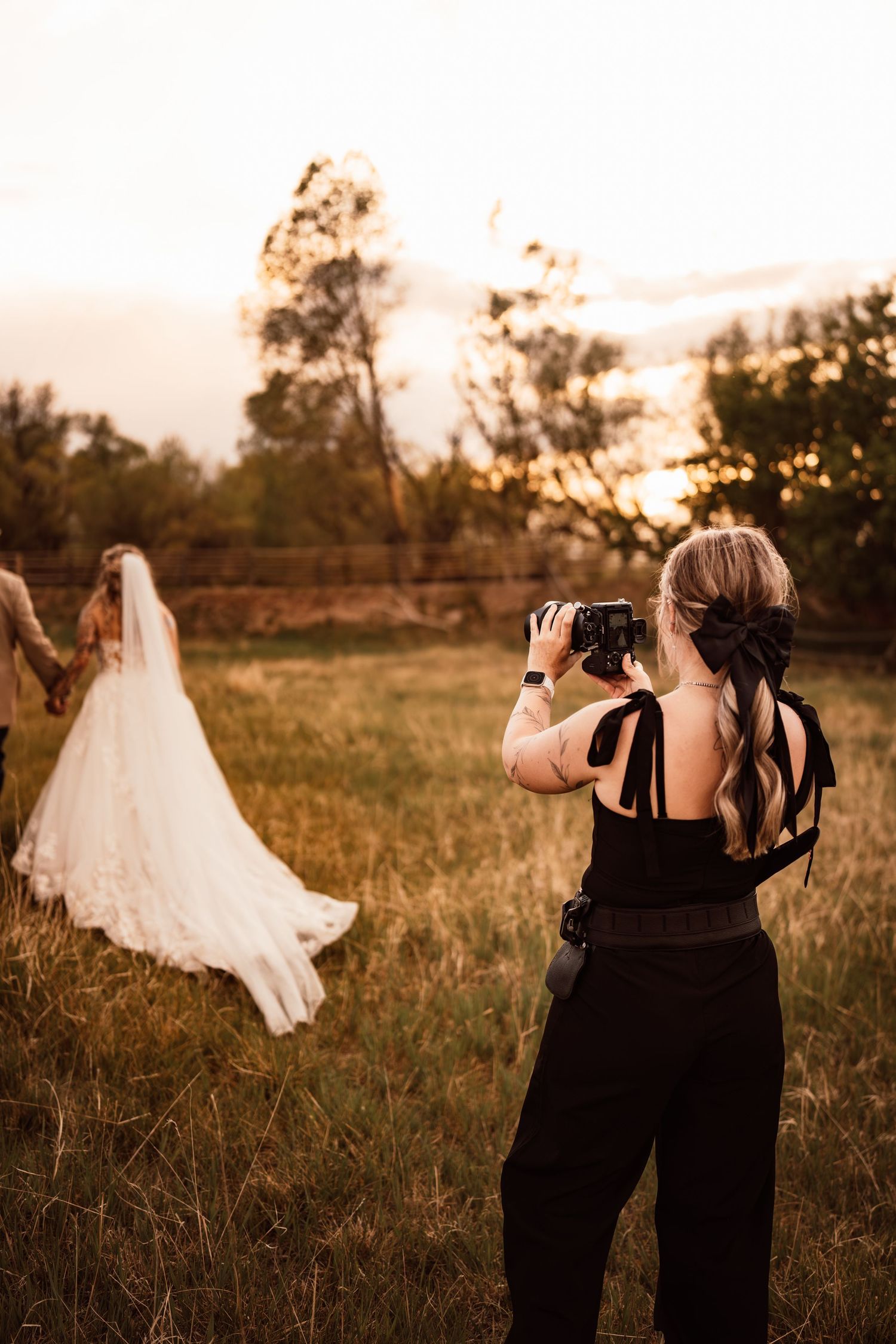 Wedding photographer in black capturing bride and groom at golden hour in a lush green field at sunset.