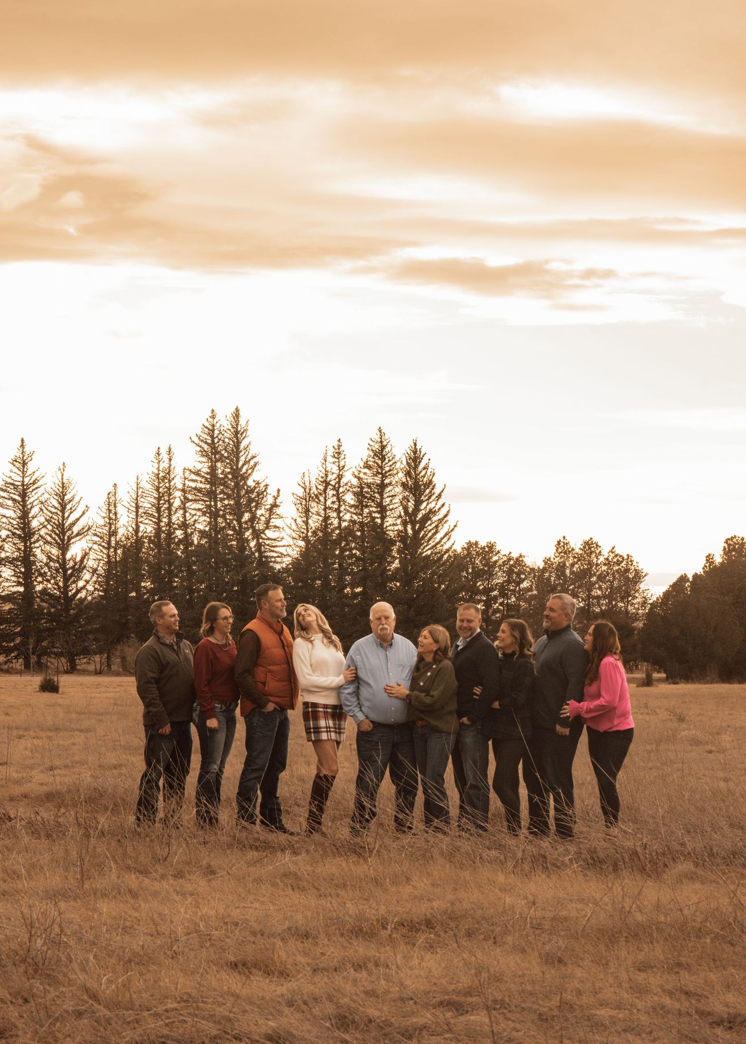 Crowd of people gathered in a golden field at sunset with pine trees in the background.