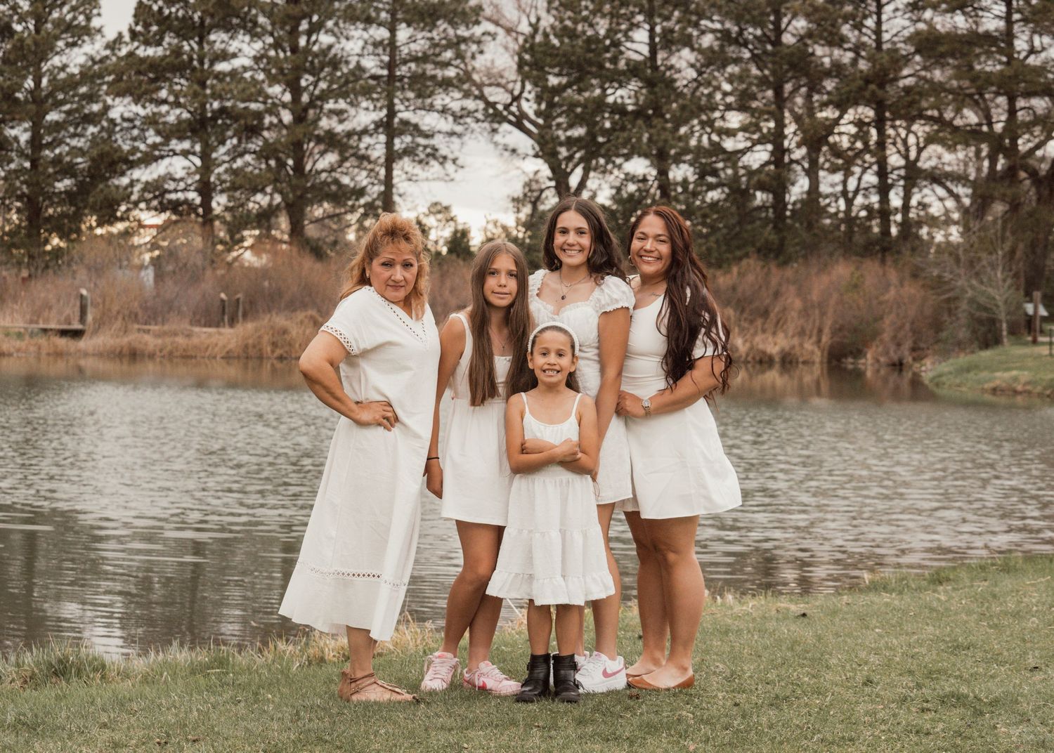 Group of women in white dresses pose together by a lake with pine trees in the background.