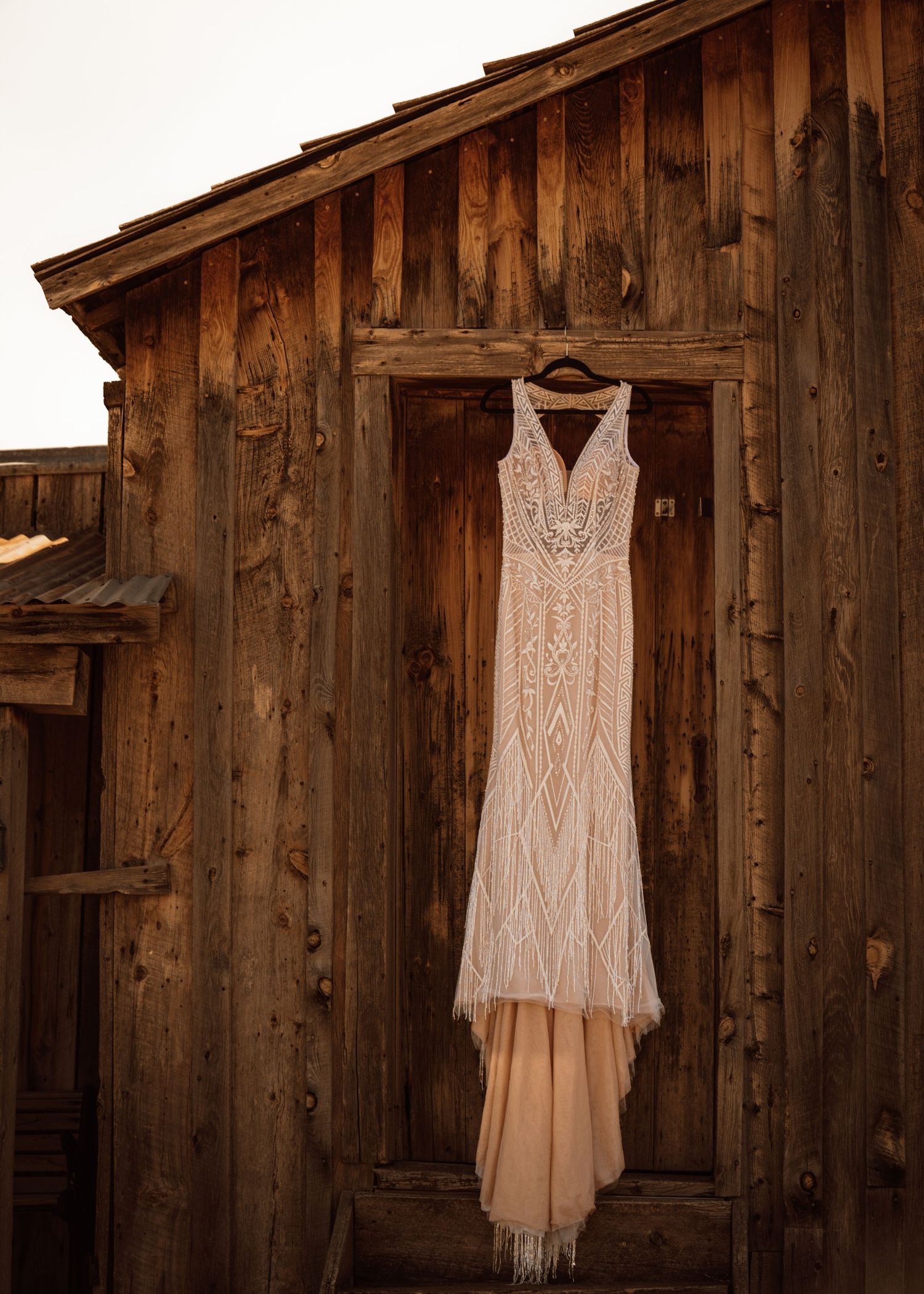 Elegant white lace wedding dress hanging on rustic wooden barn wall in natural lighting.