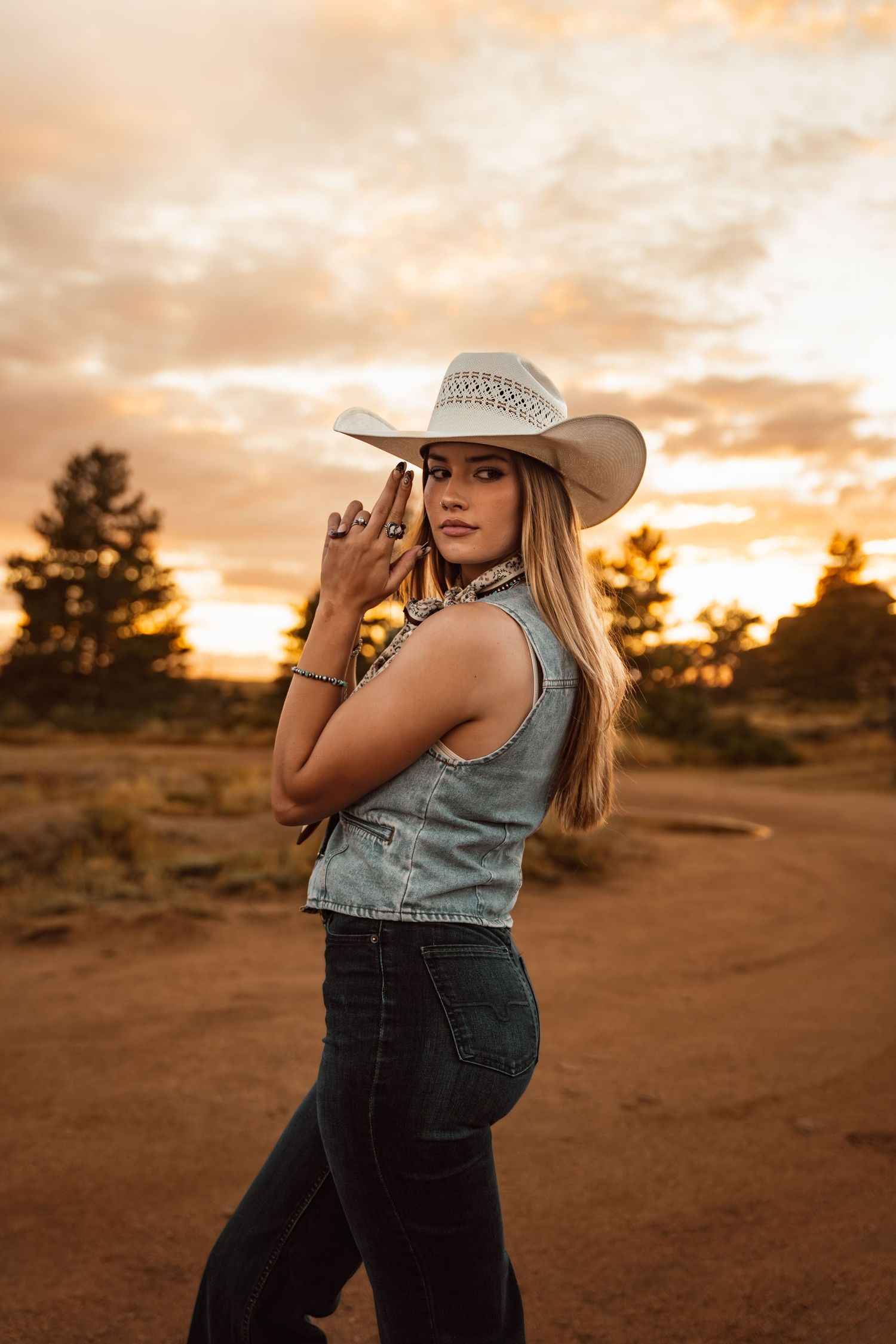 Young woman in white cowboy hat and denim vest poses confidently at golden sunset on rustic dirt road.