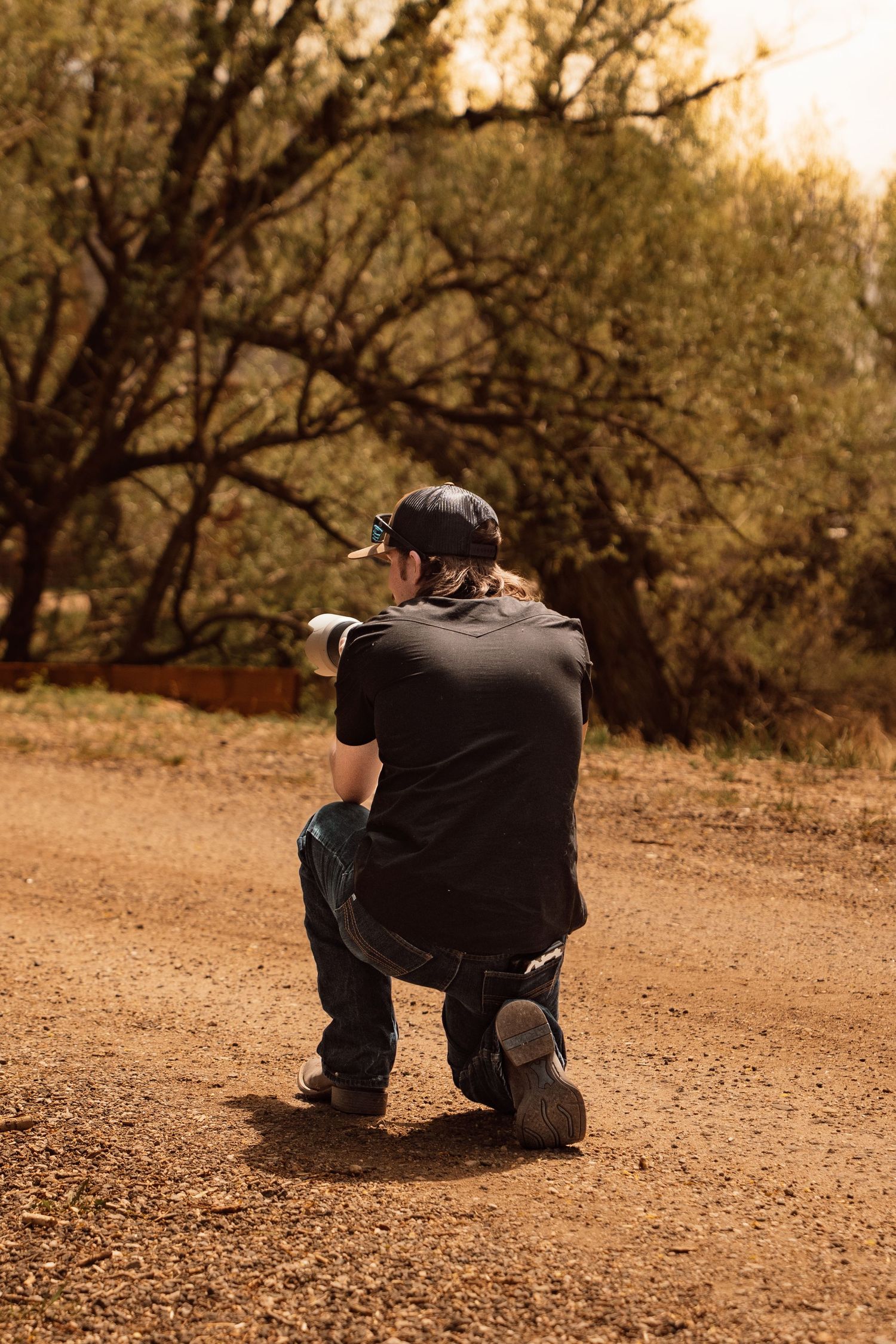 Person kneeling on a dirt path in a forest, wearing a black shirt and cap, pointing into the distance.