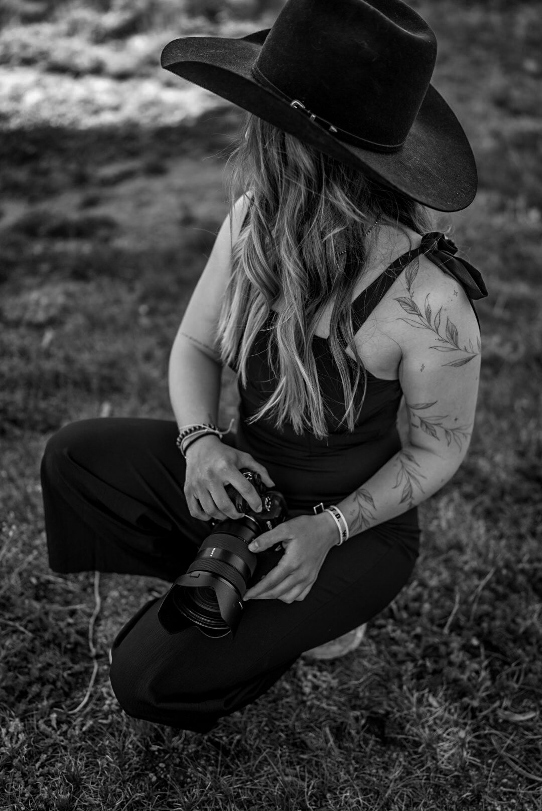 Black and white photo of a tattooed woman in a cowboy hat crouching outdoors while holding a camera.