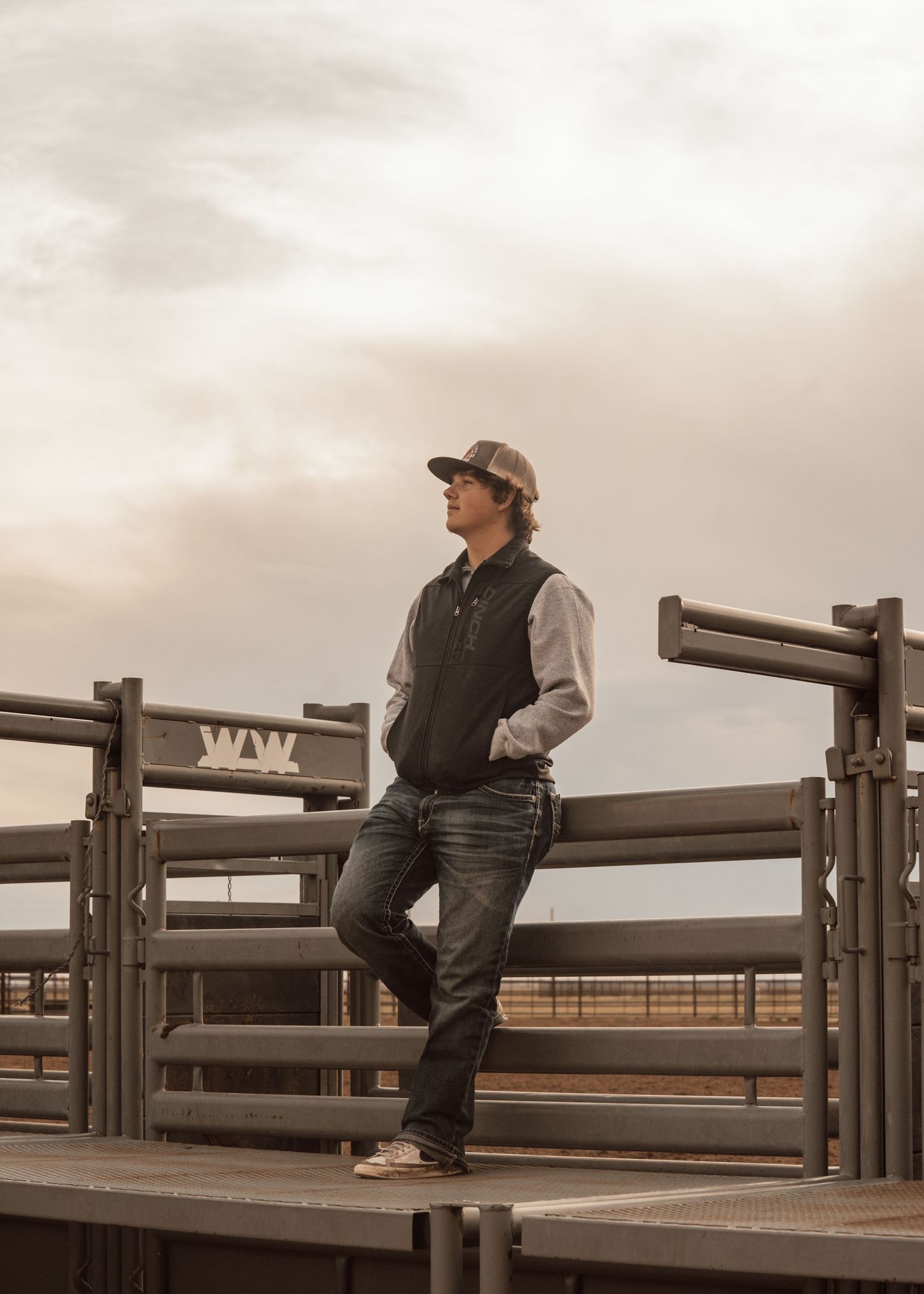 Figure in western wear poses on wooden railings at a rodeo arena during golden hour.