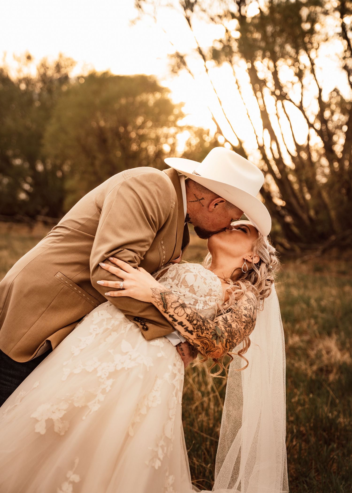 A groom in a white cowboy hat and tan suit dips his bride in a lace wedding dress for a romantic kiss outdoors.