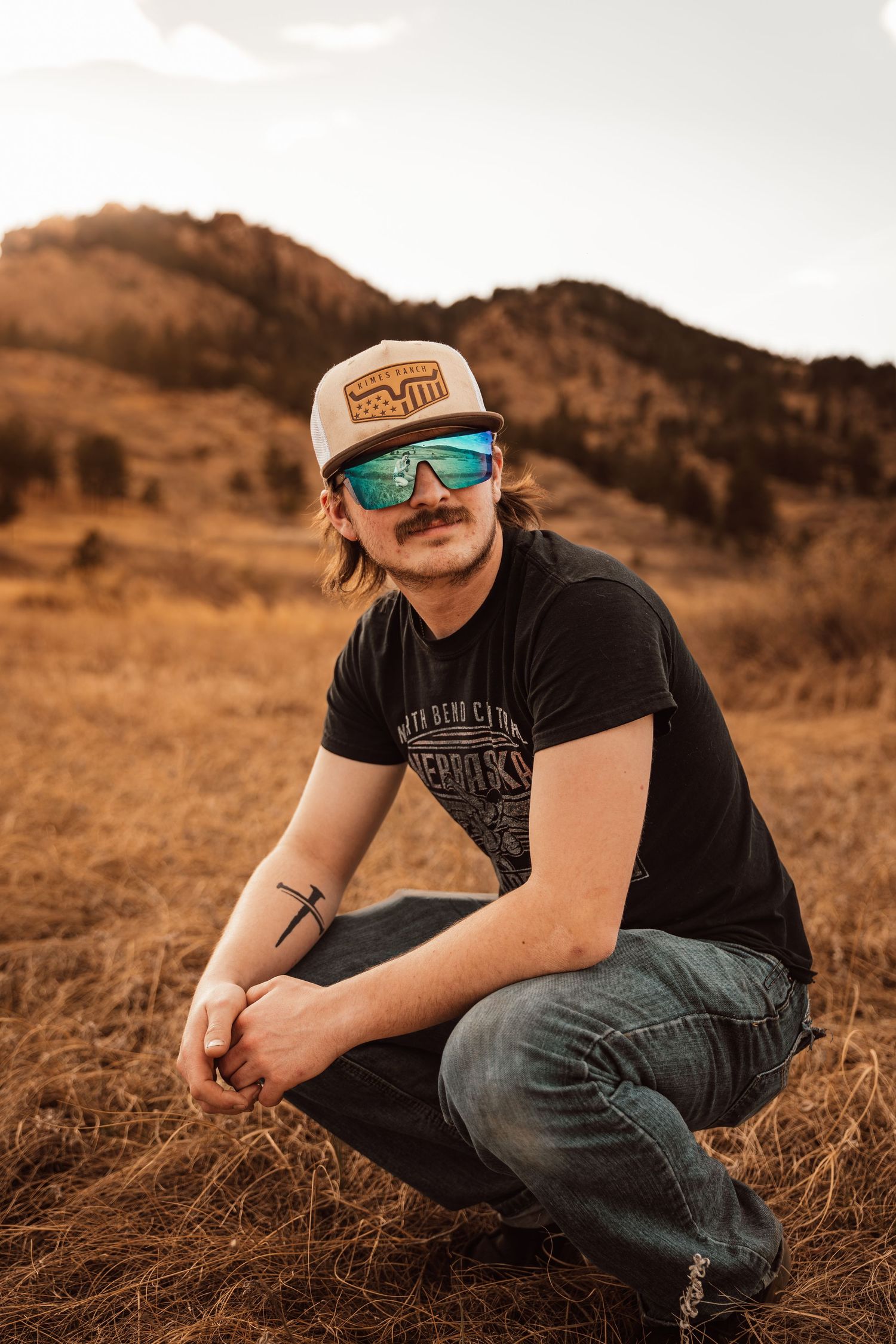 Young man with mustache wearing trucker hat and mirrored sunglasses crouching in golden field with mountains at sunset.