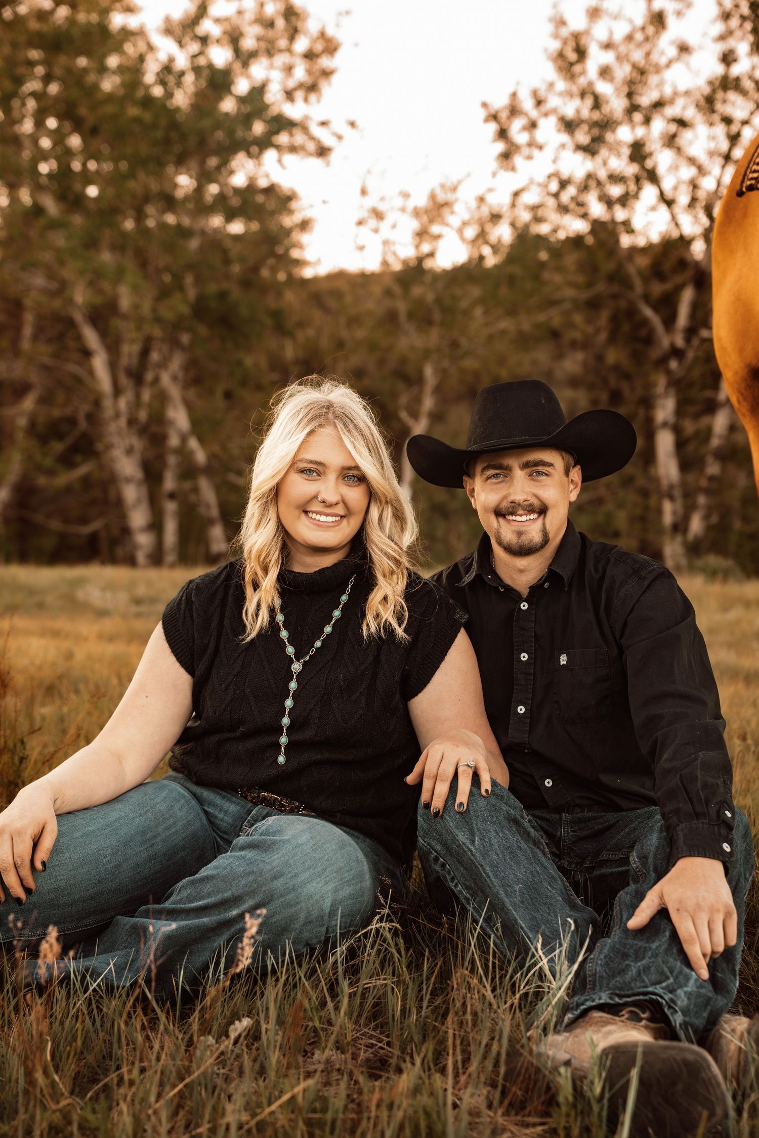 A smiling couple in matching black outfits sit together in a golden field, the man wearing a cowboy hat, with trees and a horse nearby.