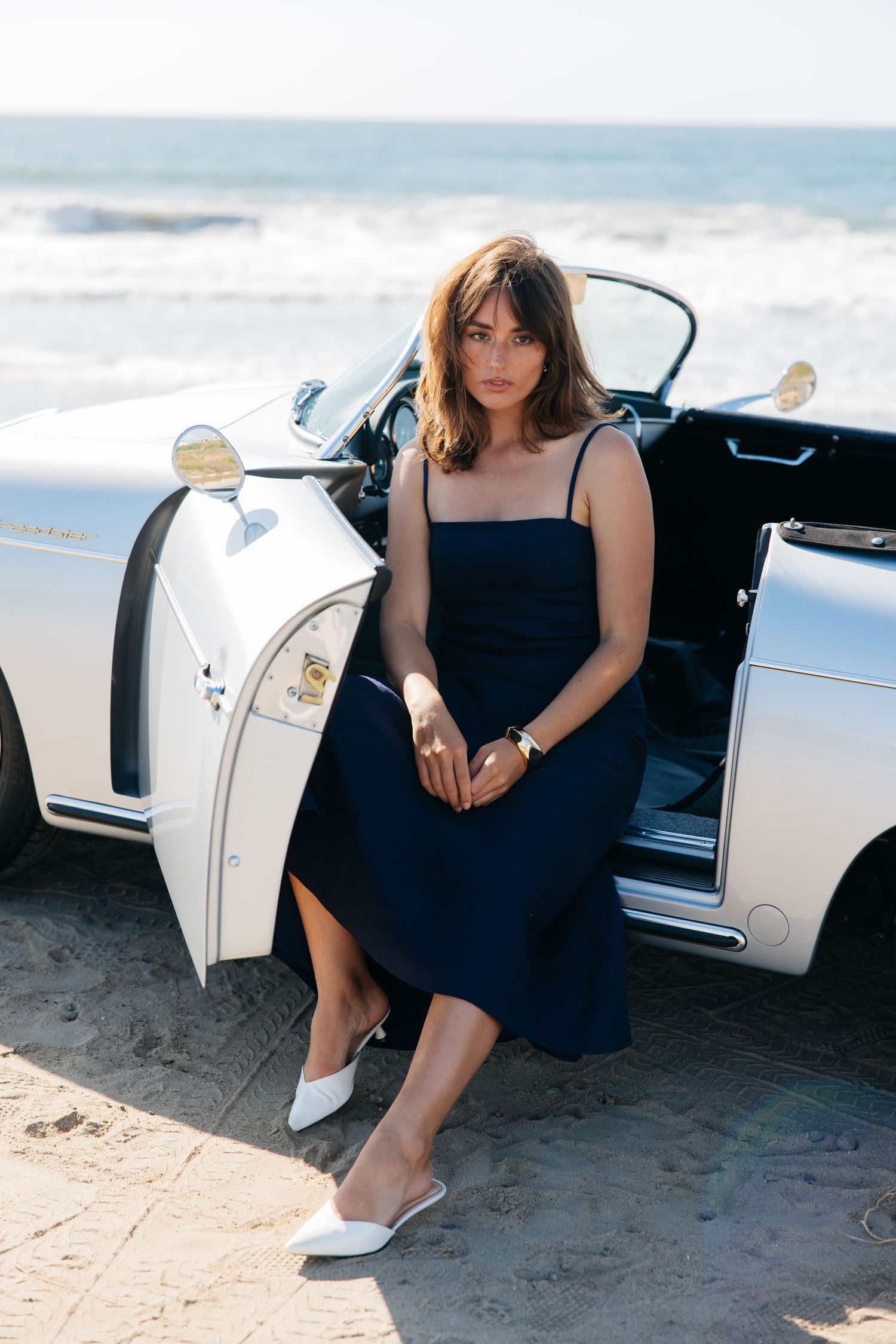 A woman in a navy blue dress sits elegantly in a white luxury car parked on a sandy beach.