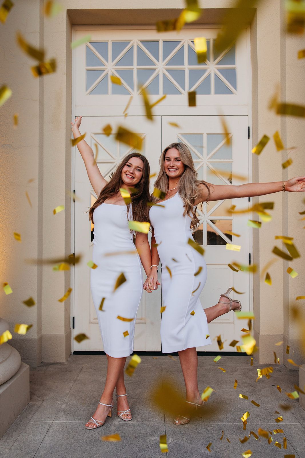 Two friends in white dresses celebrate joyfully as golden confetti falls around them in a bright hallway.