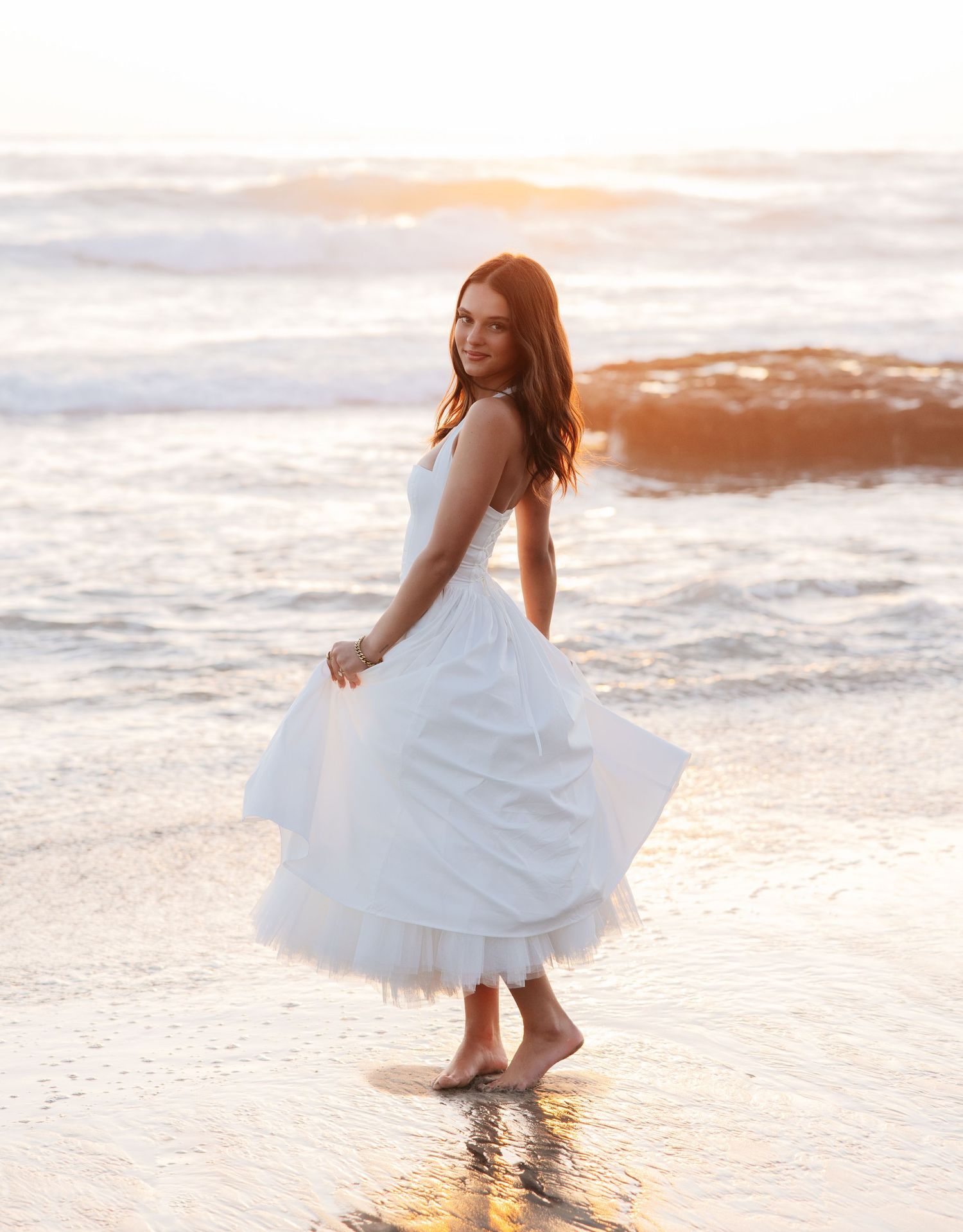 Woman in white dress twirls joyfully at sunset on a sandy beach with waves lapping at her feet.