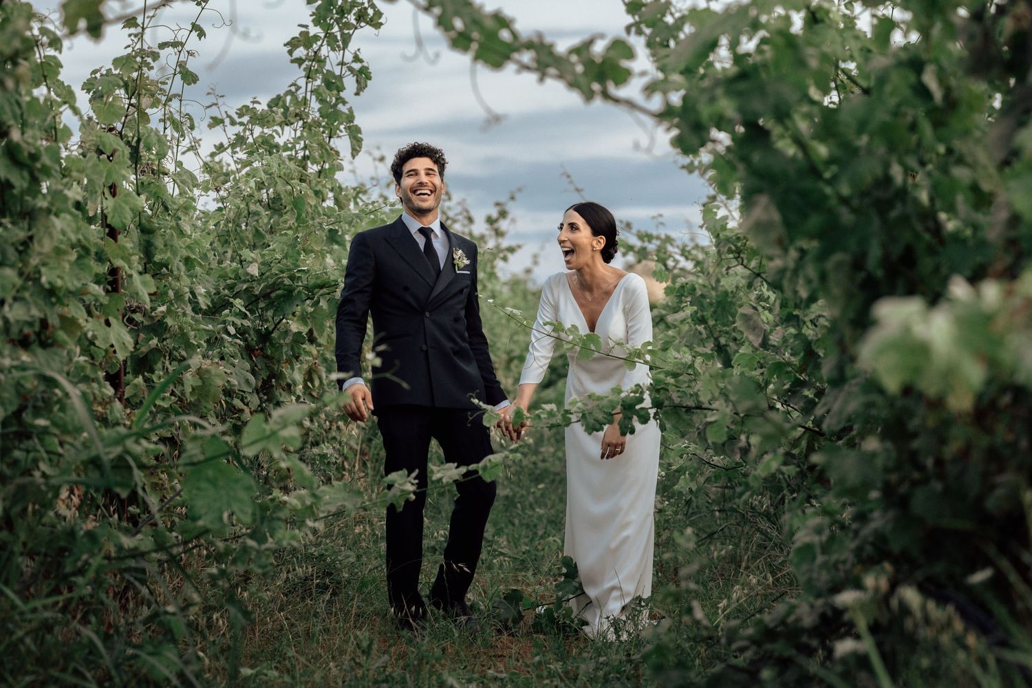 A bride and groom hold hands while standing in a lush garden pathway surrounded by green foliage.
