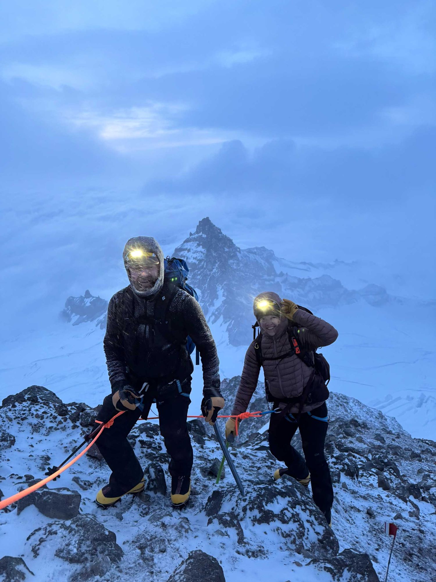Two mountaineers with headlamps climb a snowy peak in misty conditions, equipped with ice axes and safety ropes.