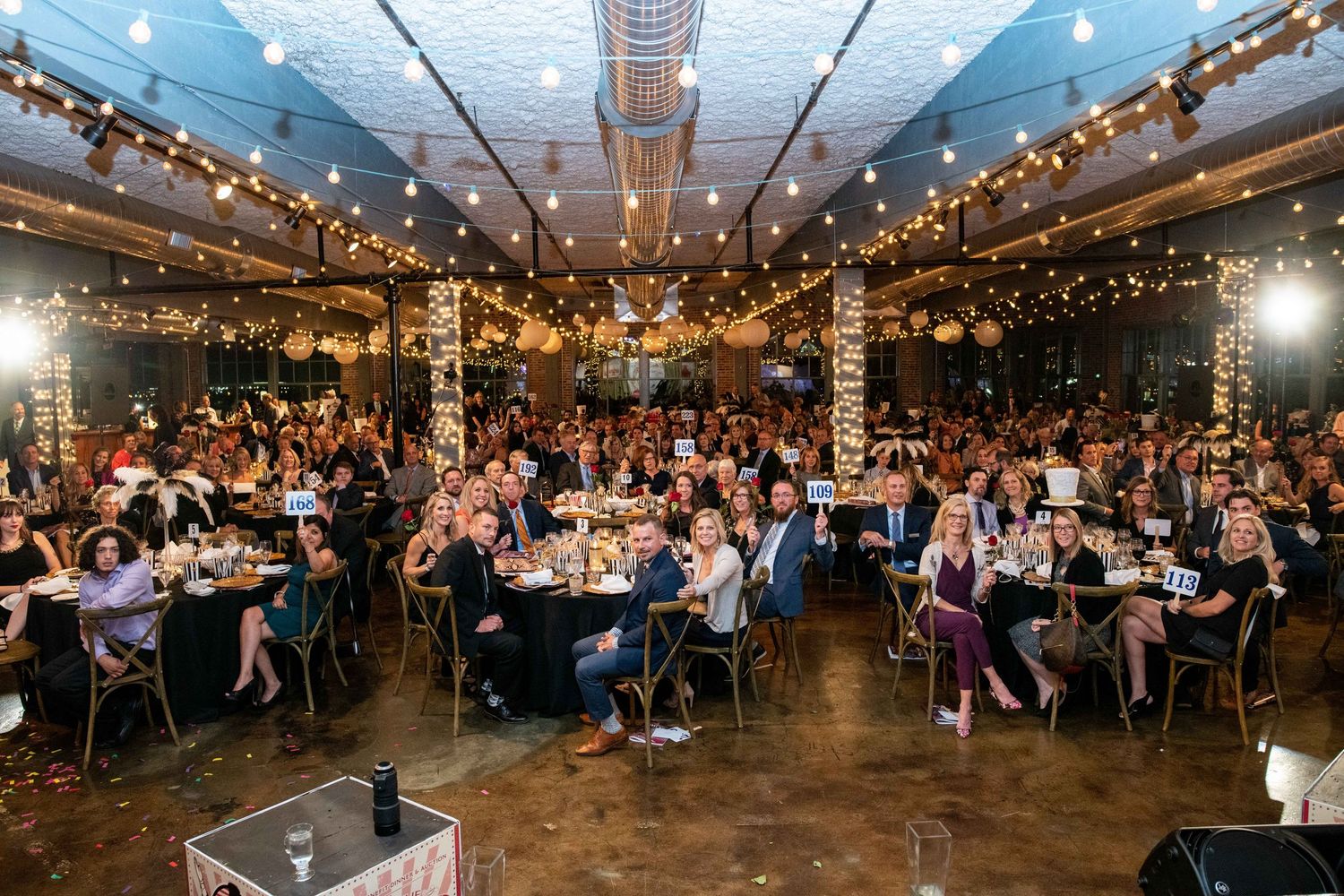Wide view of formal dinner event with string lights and exposed ceiling beams in industrial venue.