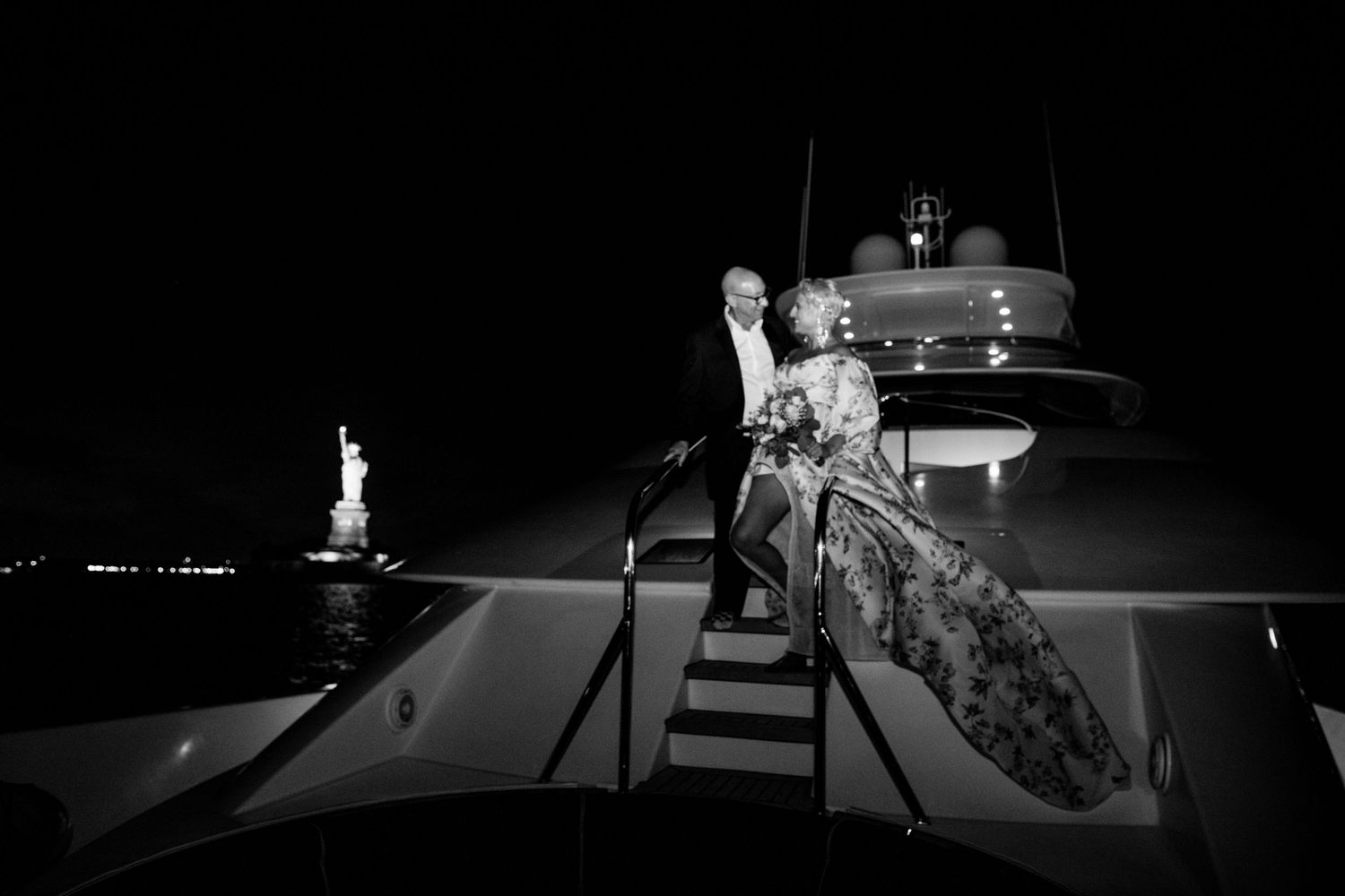 Black and white photo of couple on yacht deck with Statue of Liberty visible in distance.