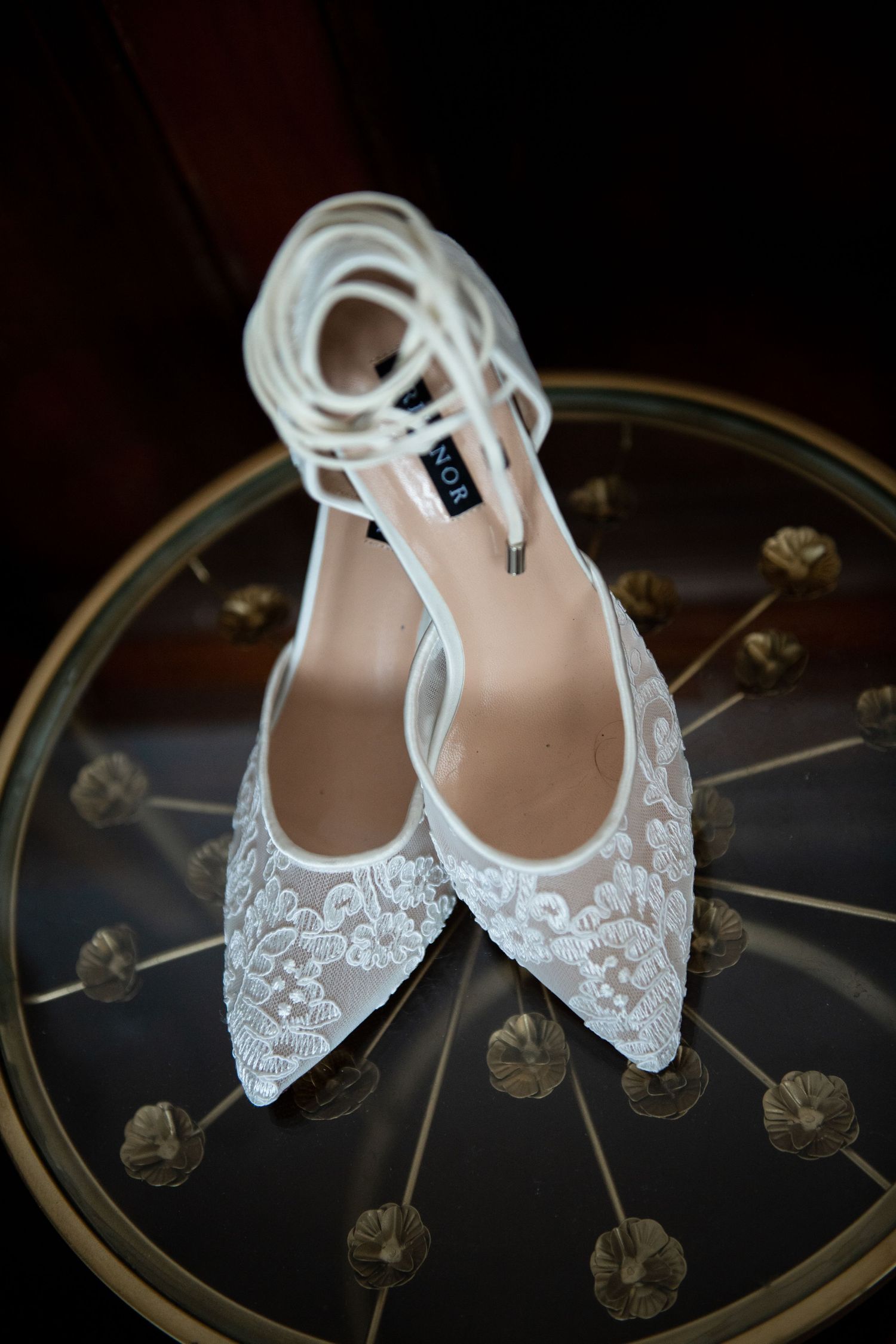 Delicate white lace bridal shoes with ankle straps displayed on dark decorative surface.