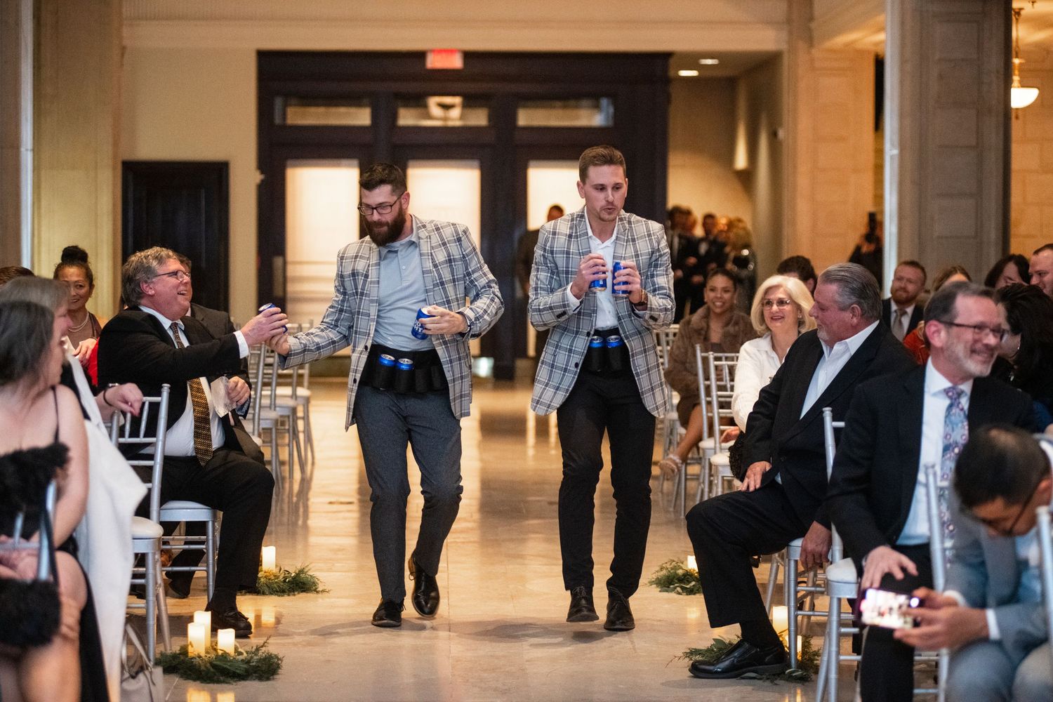 Wedding guests watch as groomsmen celebrate walking down an elegant hallway.