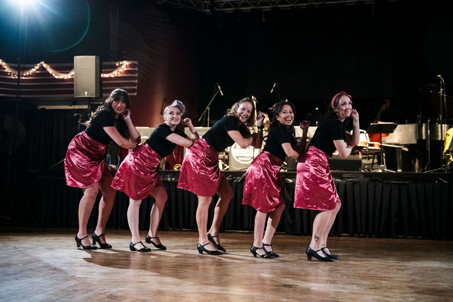 Line of dancers in matching pink dresses perform coordinated moves on a dark stage.