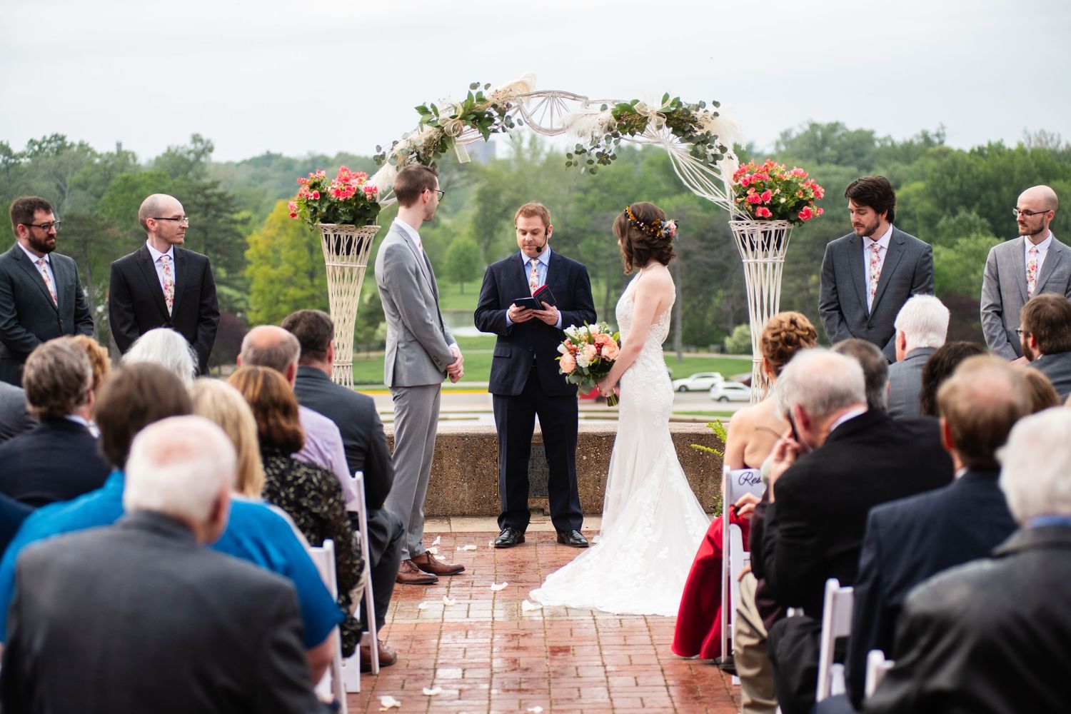 Outdoor wedding ceremony under floral arch with guests seated on lawn during sunny day.