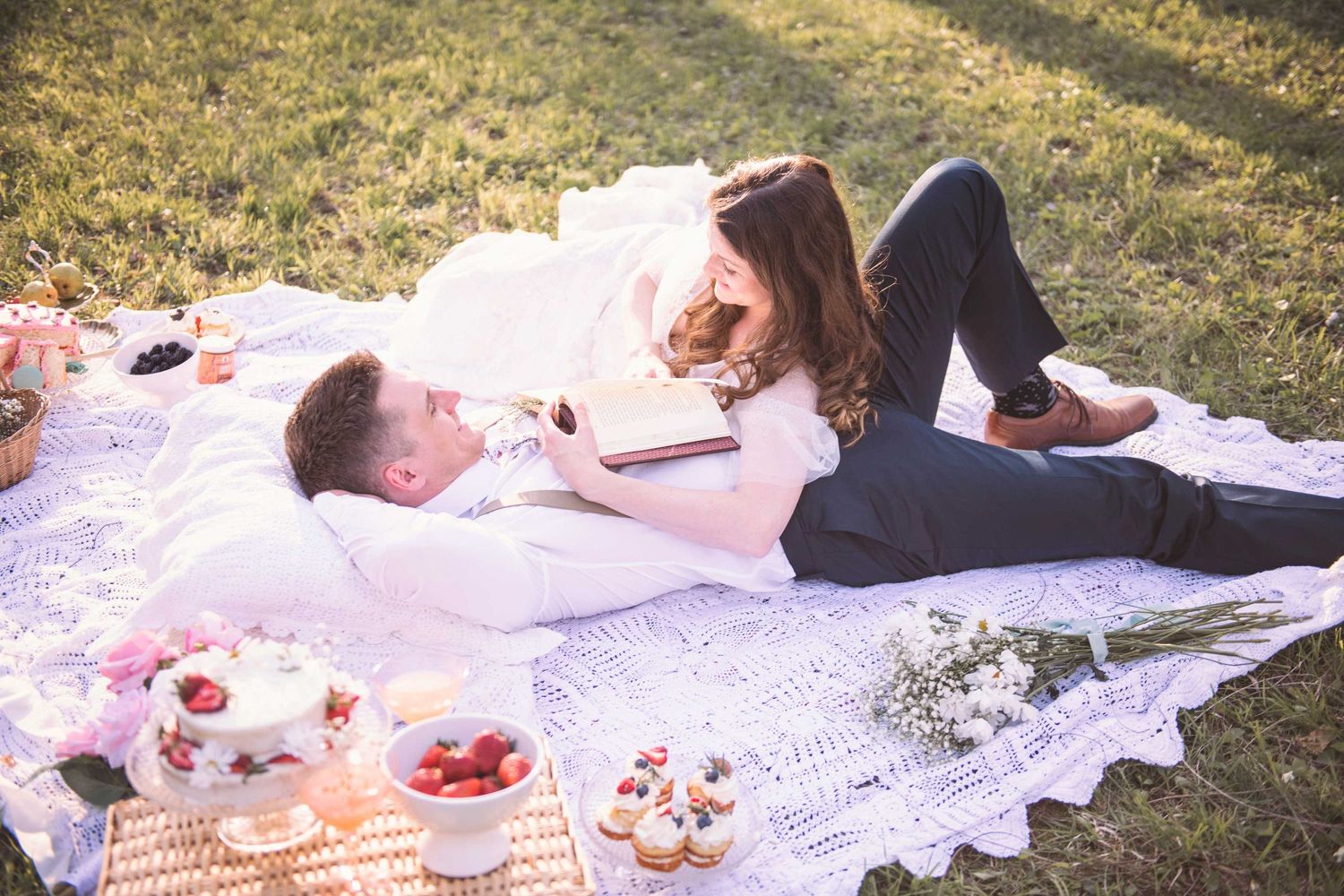 Couple enjoying a romantic picnic on a white blanket with desserts and flowers.