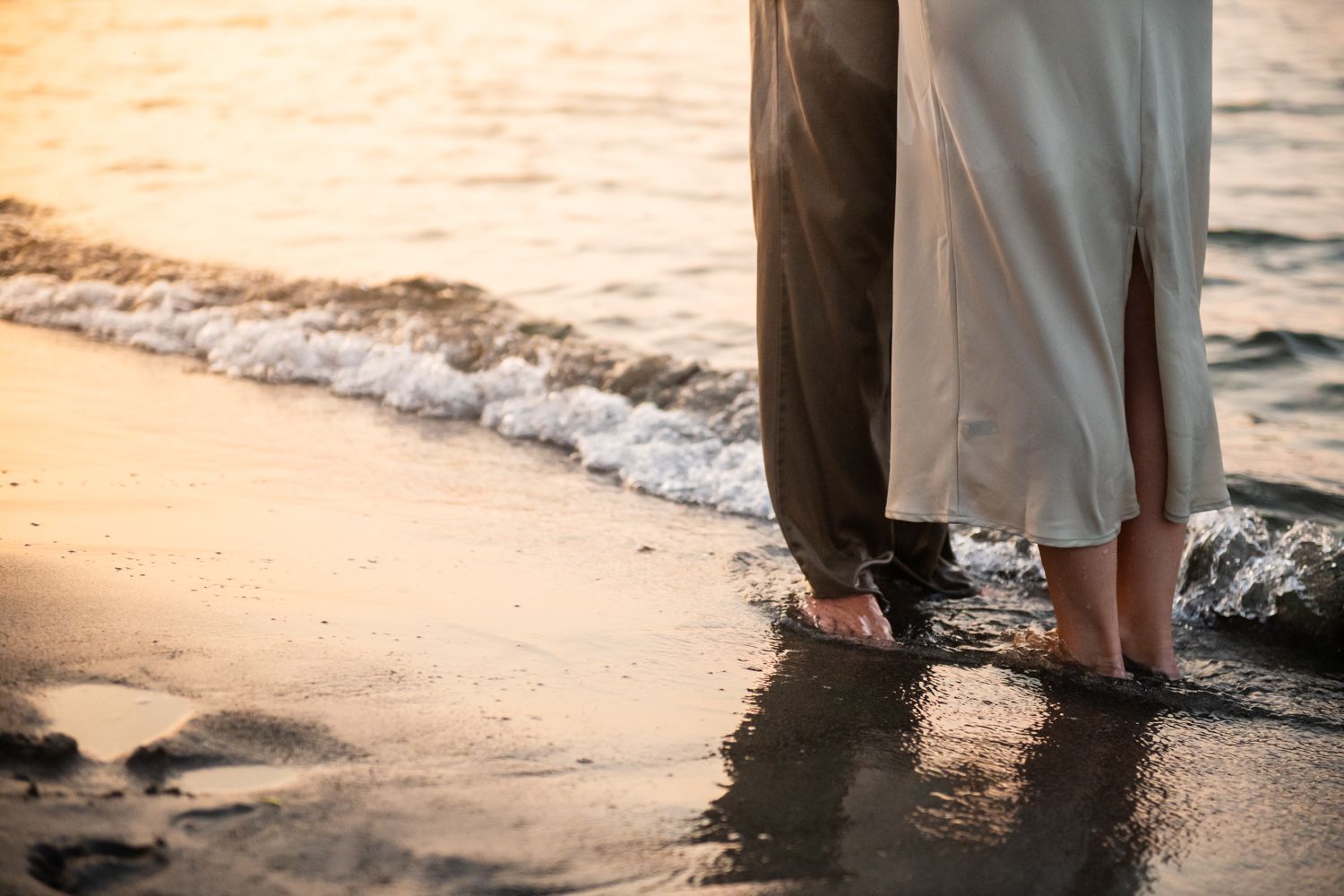 Feet of a couple standing in gentle ocean waves at sunset on a sandy beach.