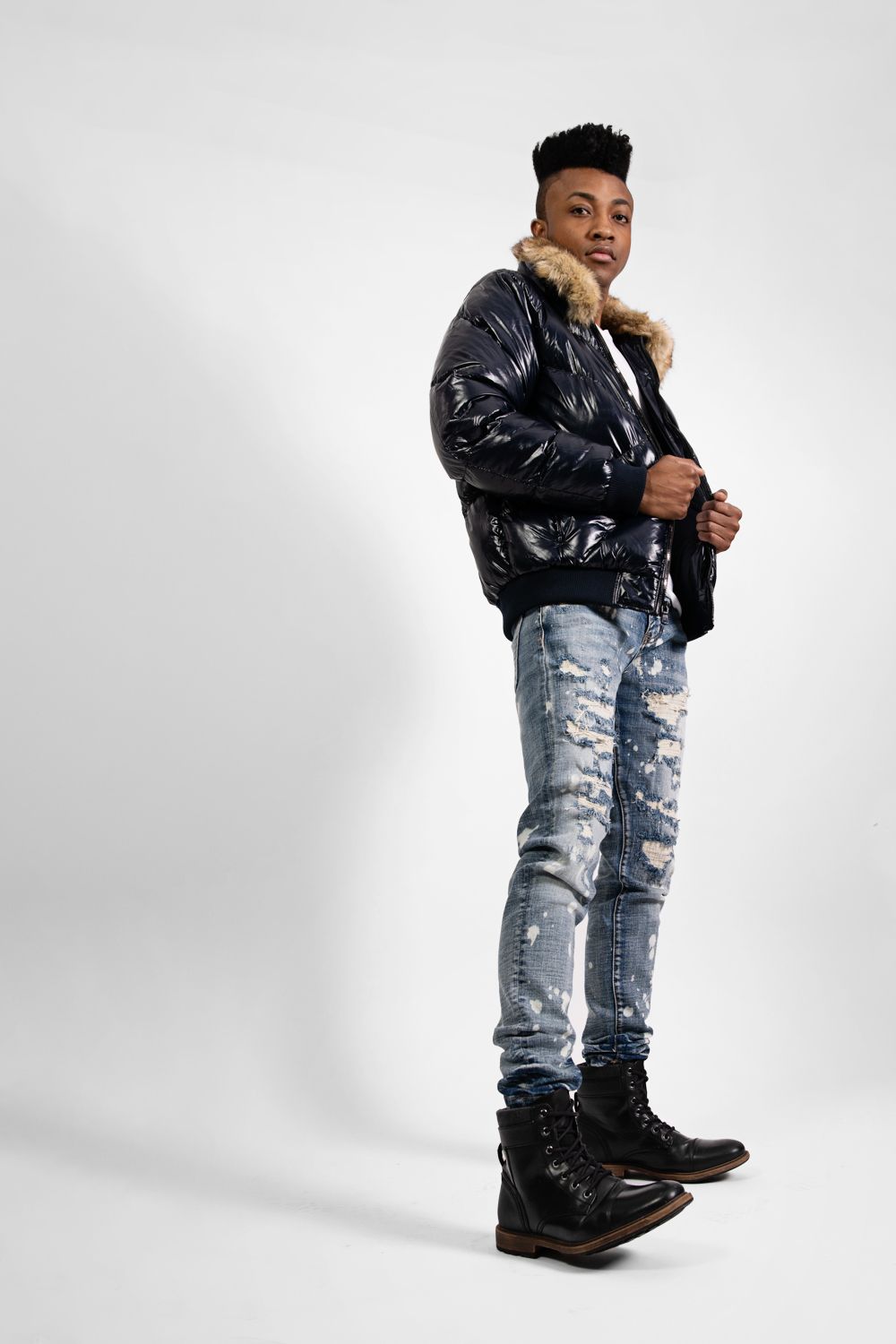 Fashionable model in shiny puffer jacket and distressed jeans poses against white backdrop.