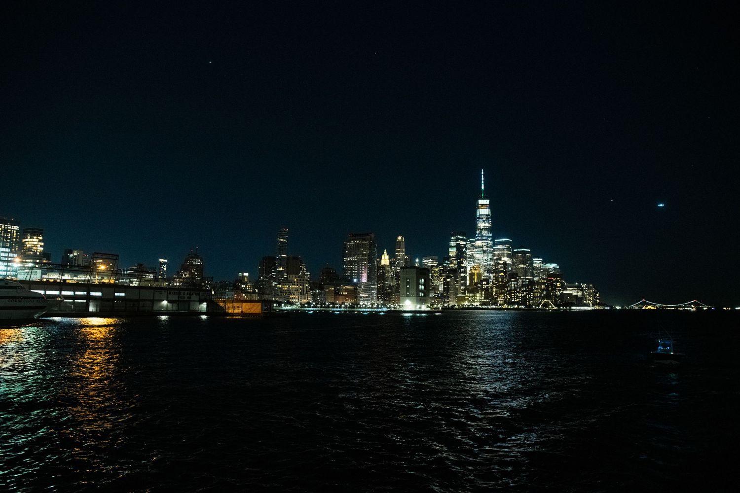 Manhattan skyline at night with Freedom Tower visible across dark waters.