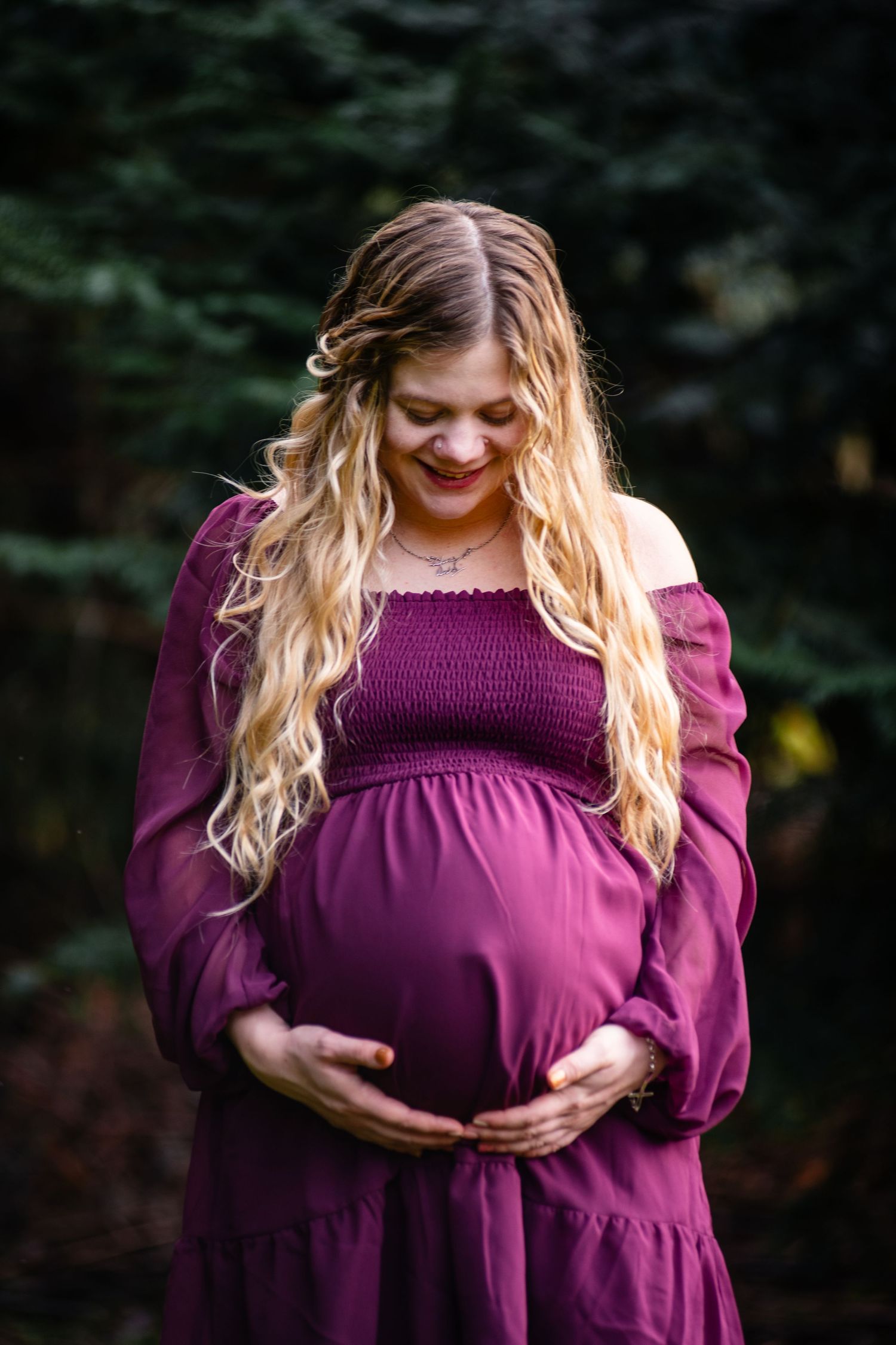 Expecting mother in a burgundy dress with long wavy blonde hair poses outdoors during maternity photoshoot.