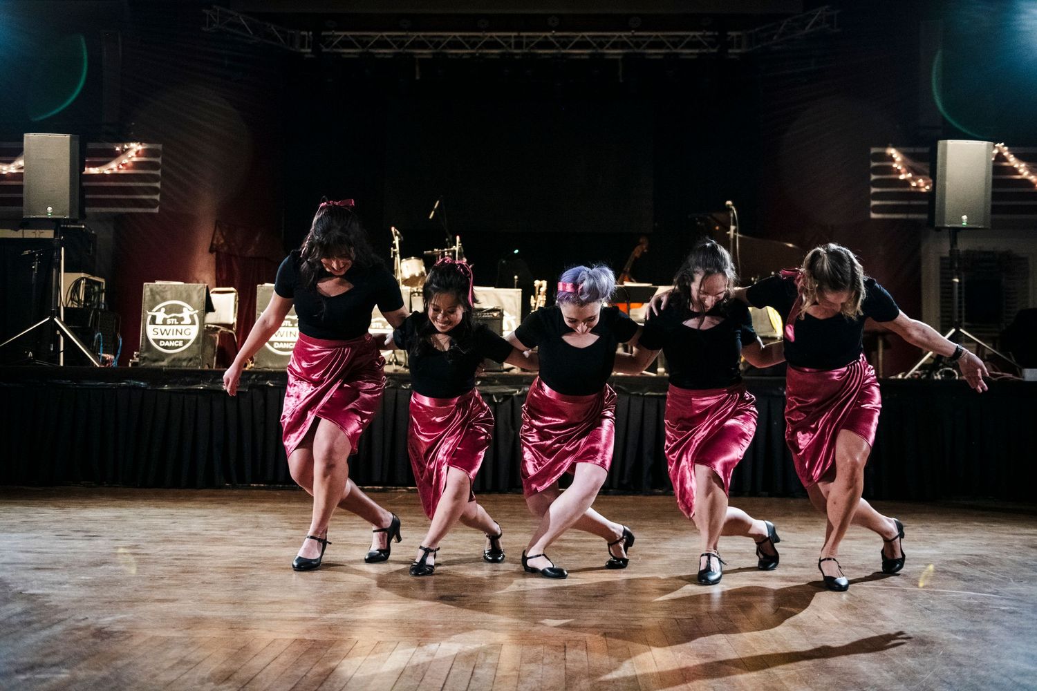 Five dancers in pink dresses perform synchronized dance moves on stage.