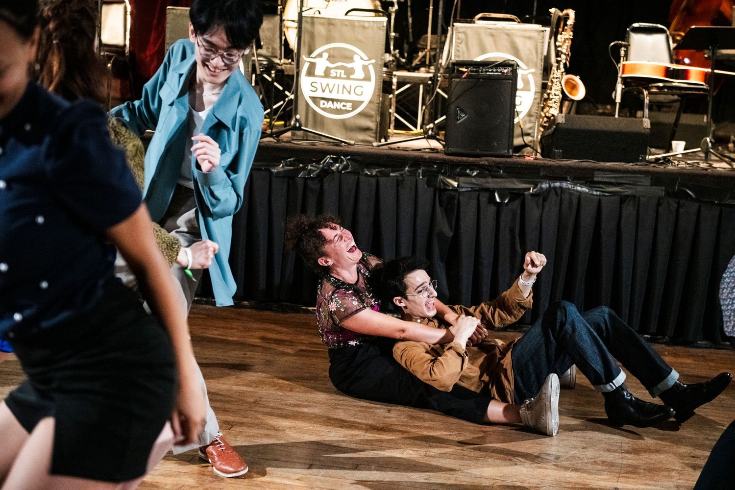 Dancers move energetically on a wooden dance floor in front of a stage with musical equipment.